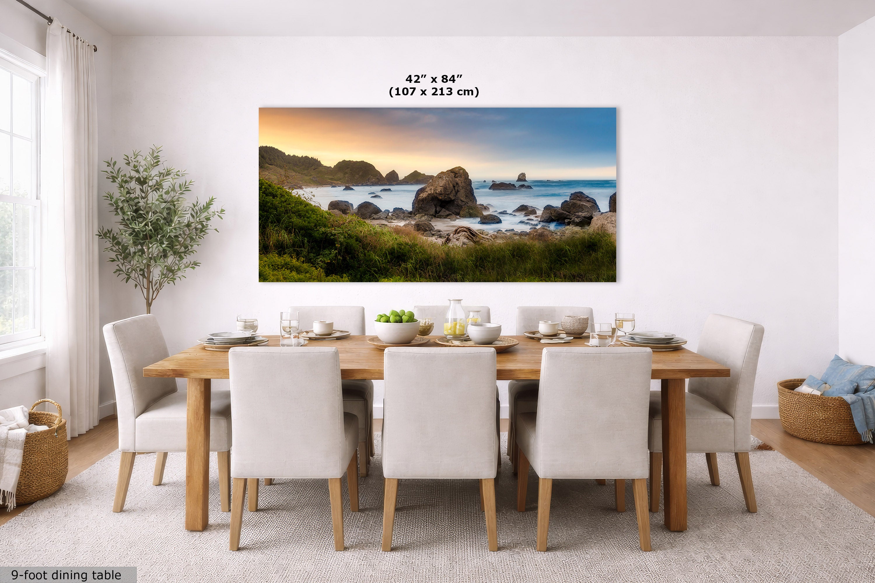 A large rectangular wall art print showing a coastal rocky beach at sunset hangs above a dining table in a bright modern room. The print includes the text '42" x 84" (107 x 213 cm)' and the scene itself is the product being sold, so this is best classified as Scenery rather than Lifestyle or Product/Packaging.