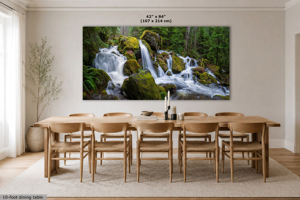 A large wall art print showing a forest waterfall scene, displayed above a wooden dining table and chairs in a bright dining room. The print includes size text reading 42 x 84 inches (107 x 214 cm) at the top, and a small note at bottom left says 10-foot dining table.