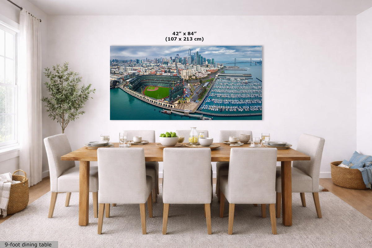 A large Oracle Park skyline panorama wall art print showing an aerial view of a harbor city and stadium, displayed above a dining table in a bright modern room. The print includes size text reading 42 x 84 inches (107 x 213 cm).