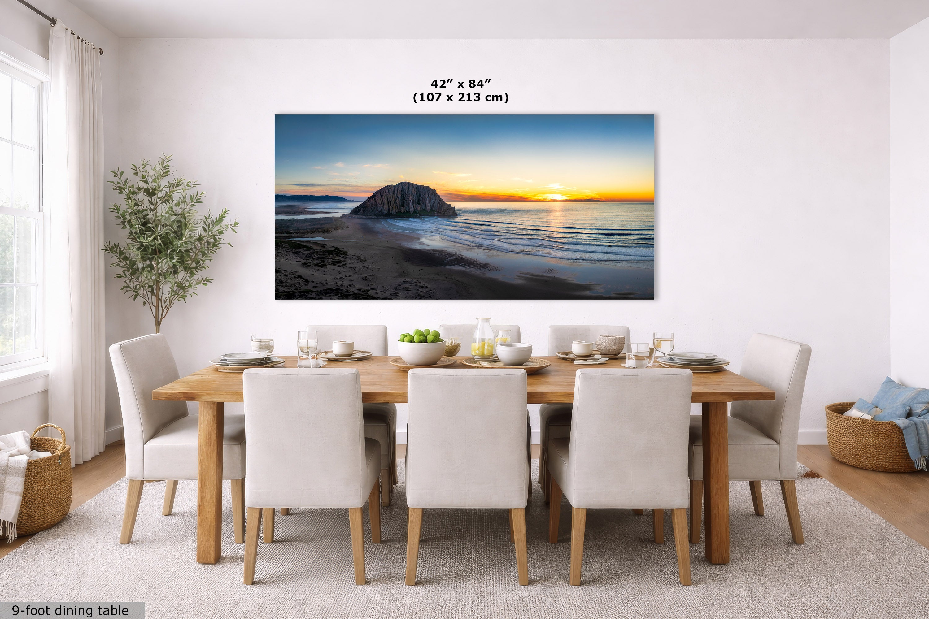 A large rectangular wall art print showing a beach and rock silhouette at sunset, displayed above a dining table in a bright modern room. Text above the print reads 42 x 84 inches (107 x 213 cm).