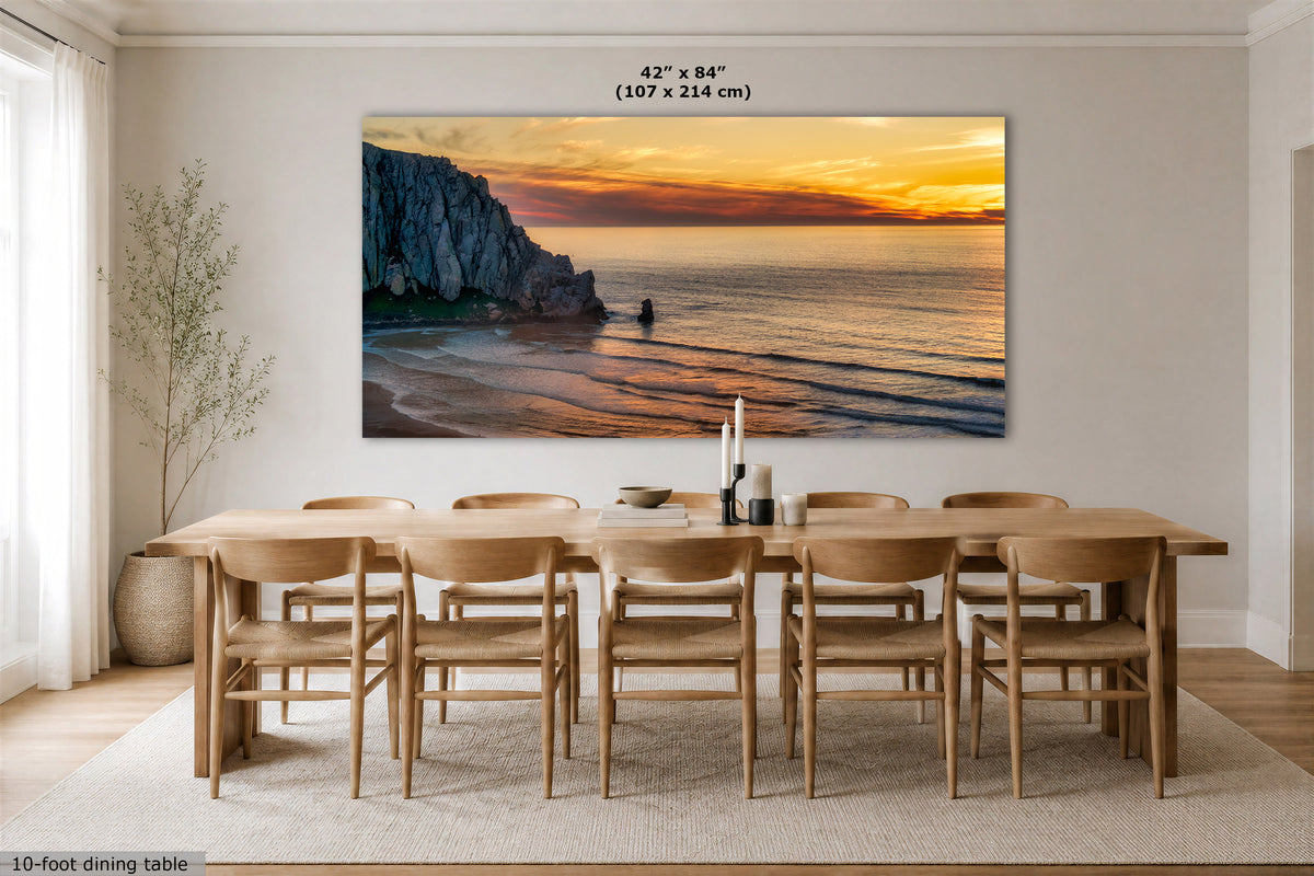A large rectangular wall art print showing a coastal cliff and ocean sunset hangs above a 10-foot dining table with ten chairs in a bright neutral dining room. The print includes size text at the top reading 42" x 84" (107 x 214 cm).