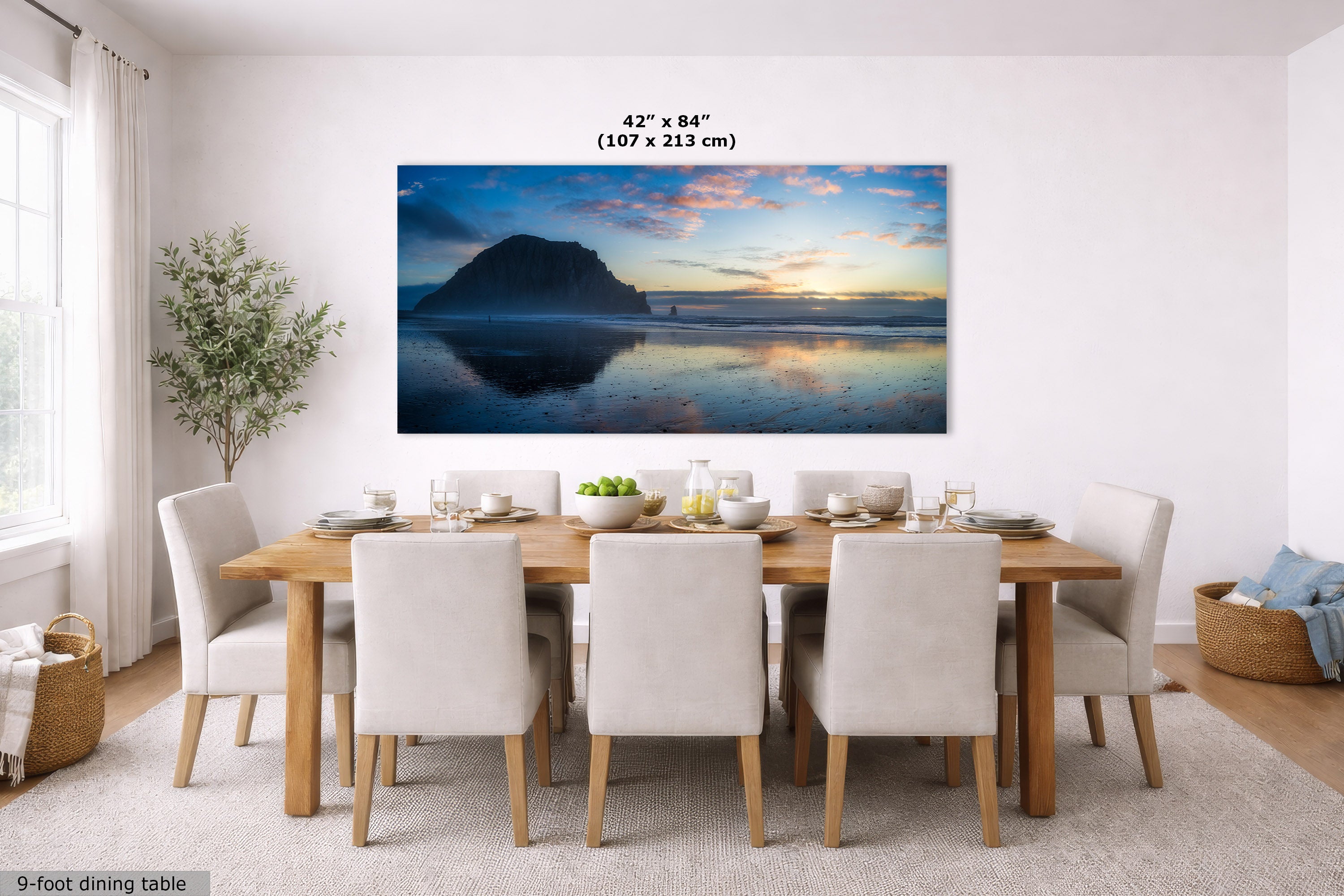 A large panoramic wall art print showing a beach and ocean sunset with a mountain silhouette, displayed above a dining table in a bright modern room. The print includes size text reading 42 x 84 inches (107 x 213 cm).