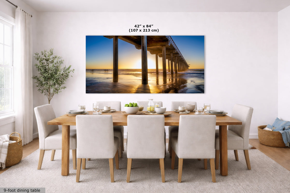 A large rectangular wall art print showing a pier over water at sunset, displayed above a dining table in a bright modern room. The print includes size text reading 42 x 84 inches (107 x 213 cm).