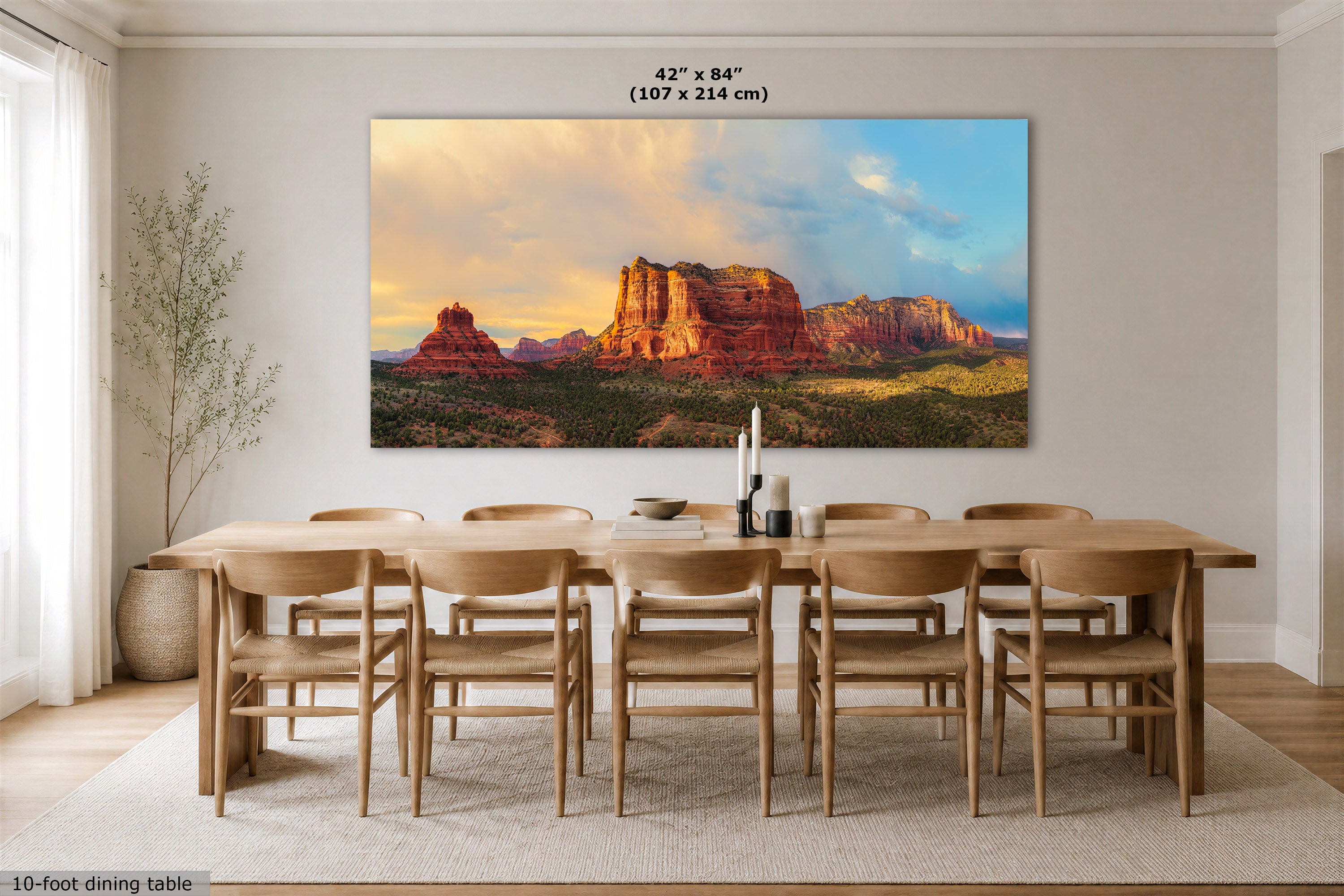 A large rectangular wall art print showing a desert rock landscape at sunset, displayed above a wooden dining table and chairs in a bright dining room. The print includes a small size label reading 42 x 84 inches (107 x 214 cm).