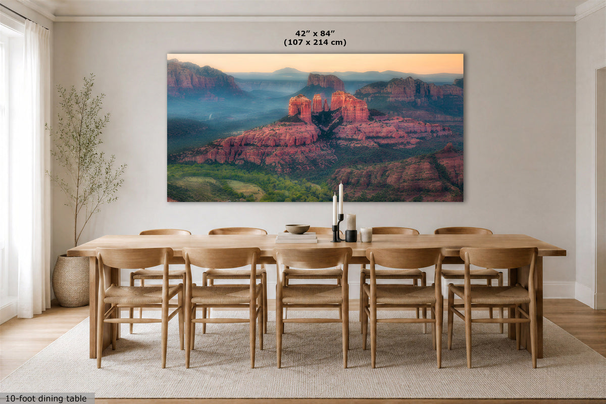 A large rectangular wall art print showing a desert canyon landscape with red rock formations and blue mist hangs above a wooden dining table and chairs in a bright dining room. Text at the top reads 42 x 84 inches (107 x 214 cm), and text at the bottom left says 10-foot dining table.