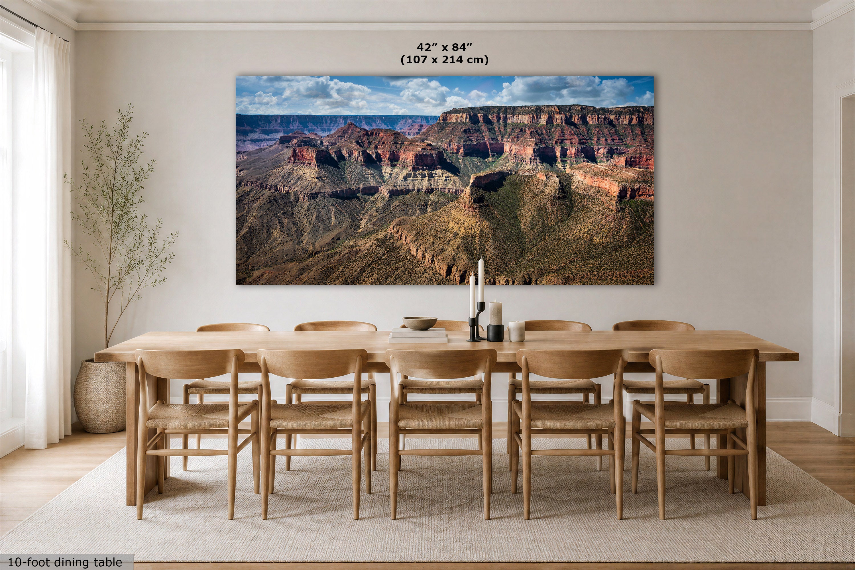A large rectangular wall art print showing a Grand Canyon landscape hangs above a wooden dining table and chairs in a bright, modern dining room. The print includes size text at the top reading 42" x 84" (107 x 214 cm).