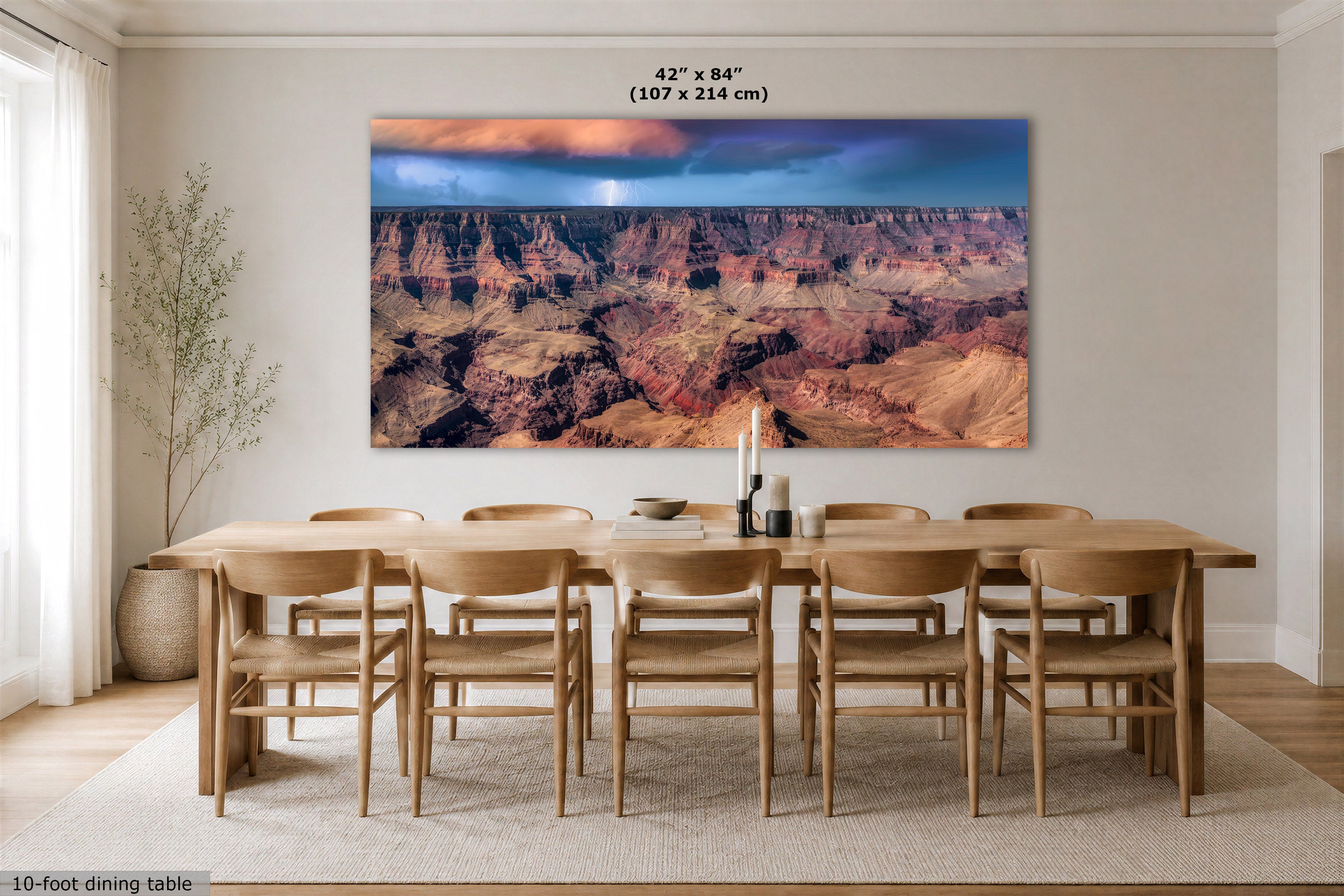 A large rectangular wall art print showing the Grand Canyon is mounted above a wooden dining table in a bright dining room. The print depicts layered canyon rock in red, brown, and tan tones under a blue and orange sky.