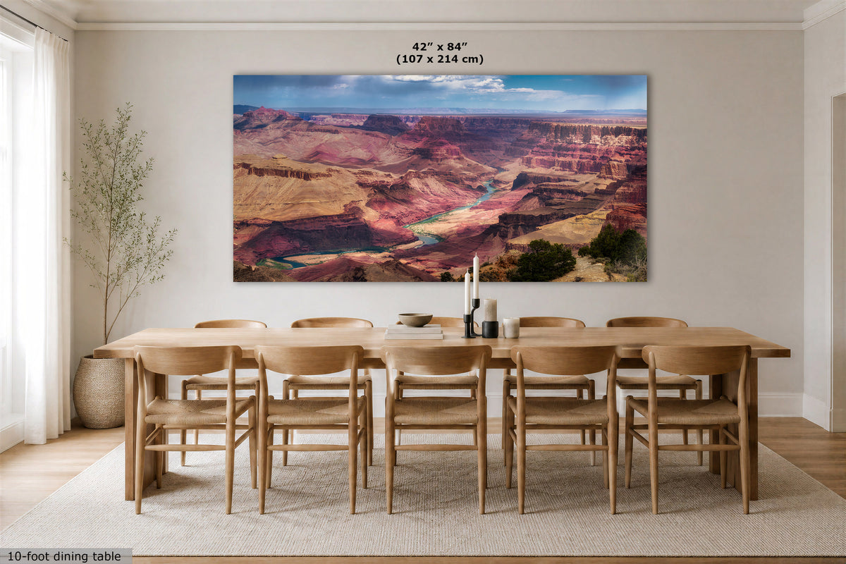 A large rectangular wall art print showing the Grand Canyon landscape hangs above a 10-foot dining table and chairs in a bright, modern dining room. The print includes dimension text at the top reading 42" x 84" (107 x 214 cm).