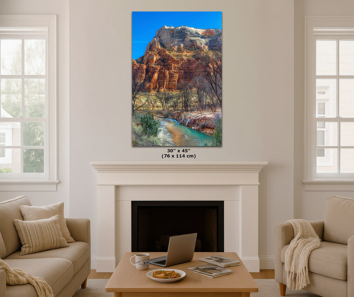 A vertical canvas print of a red rock canyon landscape with a river is displayed on a white wall above a fireplace in a modern living room setting. The print features a blue sky, orange-brown cliffs, and green water.