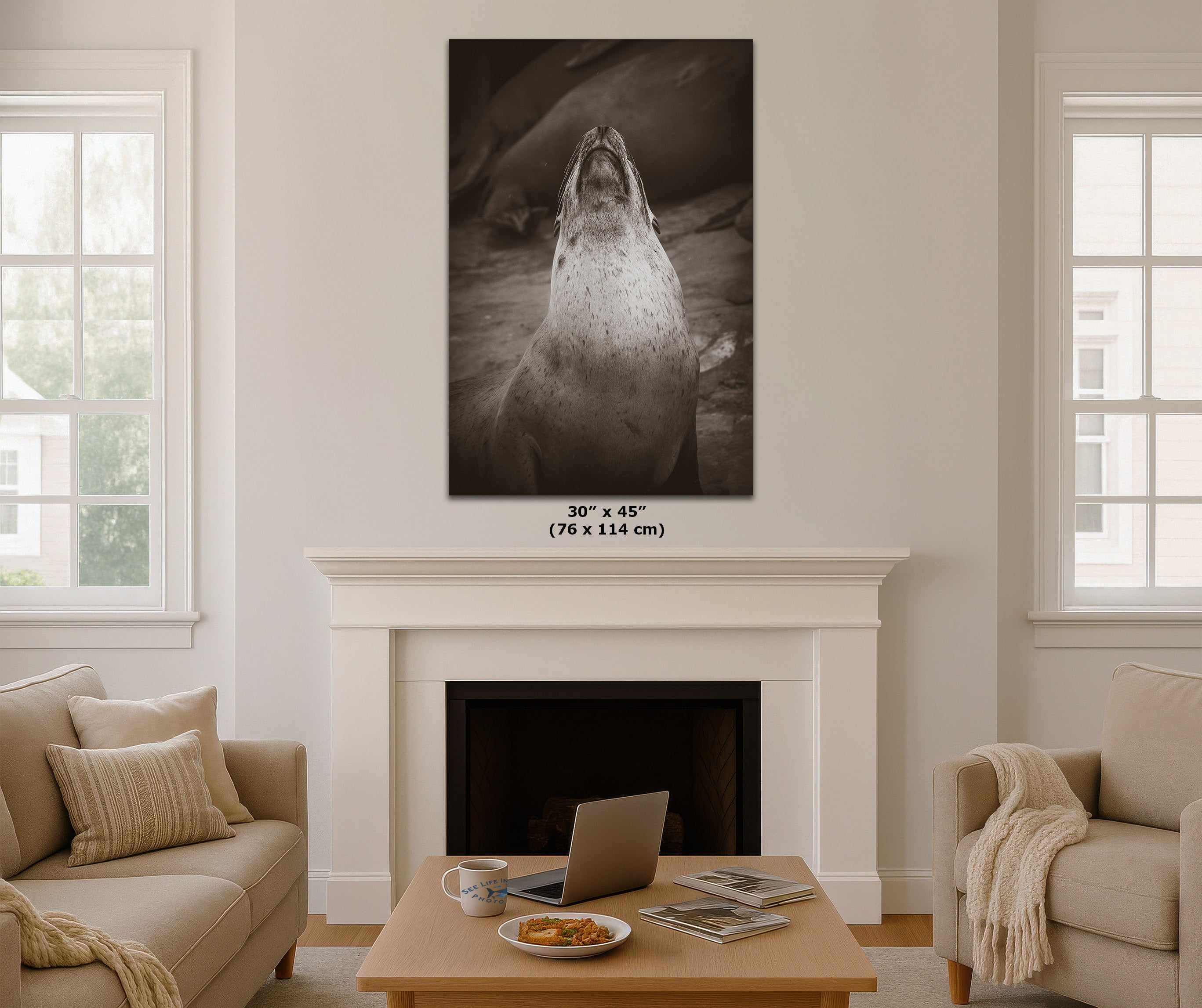 A large black and white canvas wall art print of a sea lion is displayed in a living room mockup above a white fireplace mantel. The room features beige sofas, a wooden coffee table with a laptop, and two windows on either side.