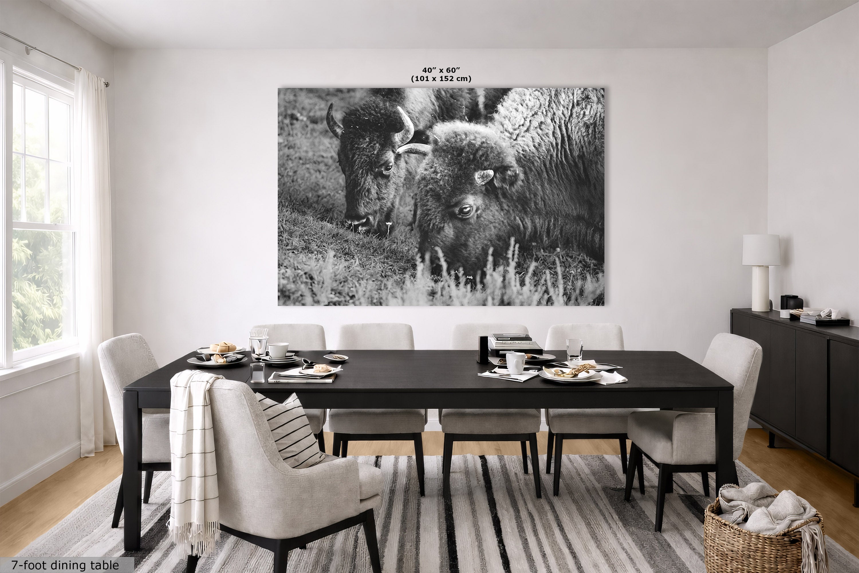 A large black and white wall art print depicting two bison in a grassy field, displayed in a modern dining room setting above a dark wood dining table and chairs. The print has dimensions labeled at the top as 40" x 60" (101 cm x 152 cm).