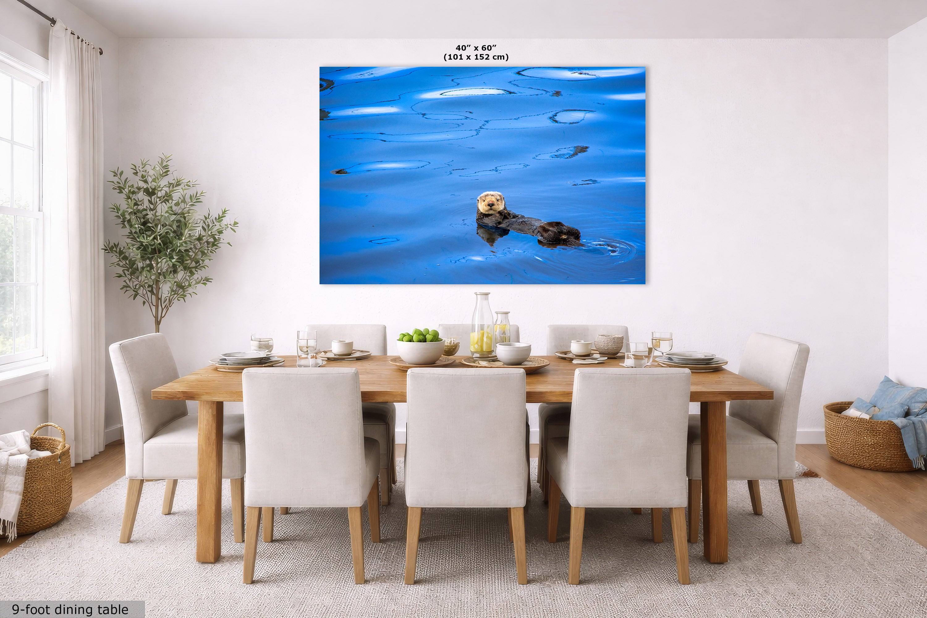 A large wall art print showing a sea otter floating on blue water, displayed above a wooden dining table with white upholstered chairs in a bright modern dining room. The print includes size text at the top and a small label at bottom left.