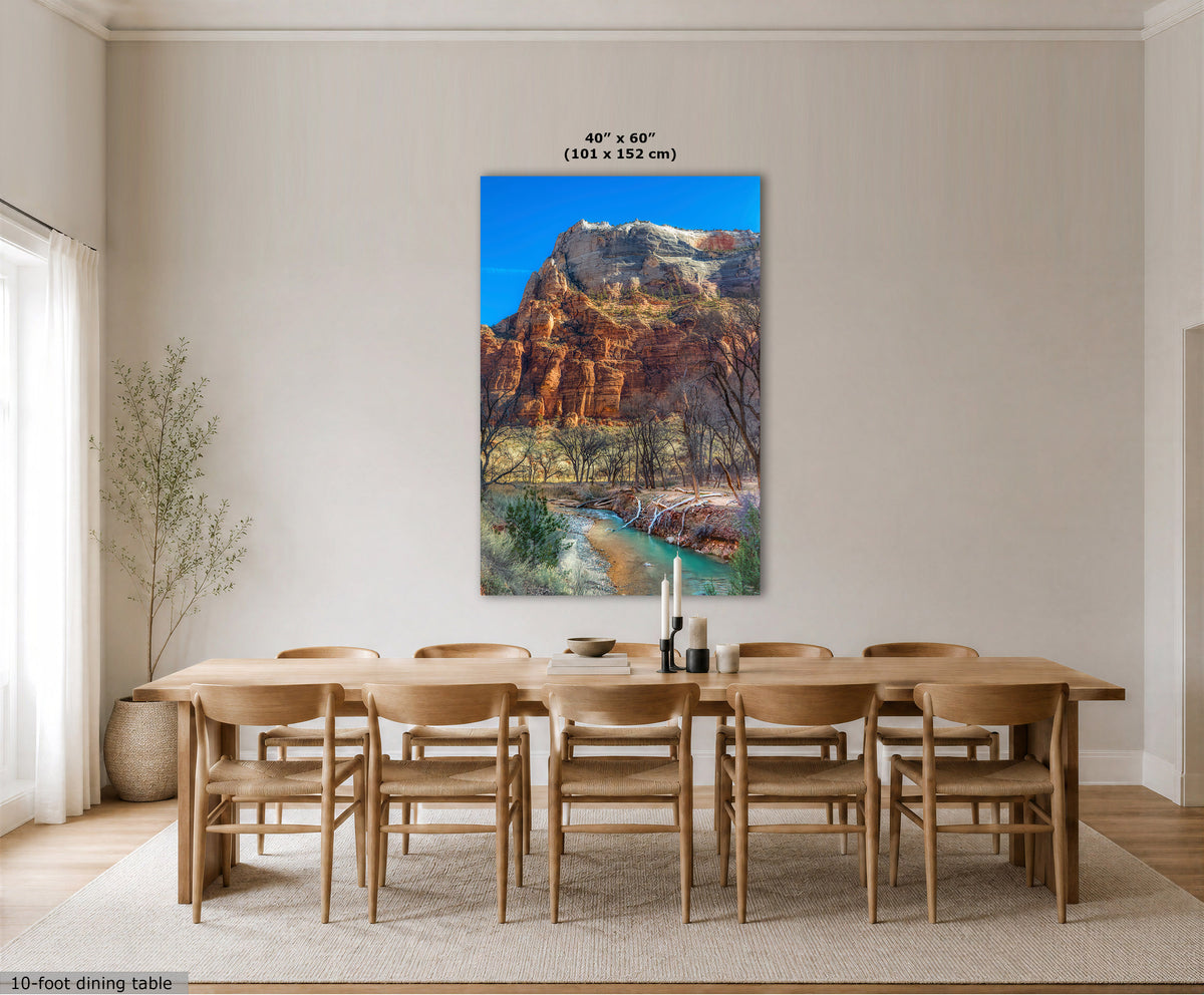 A large vertical canvas print of a desert canyon landscape with a river is displayed on a dining room wall above a wooden dining table and chairs. The room has beige walls, a light rug, and a small potted plant.