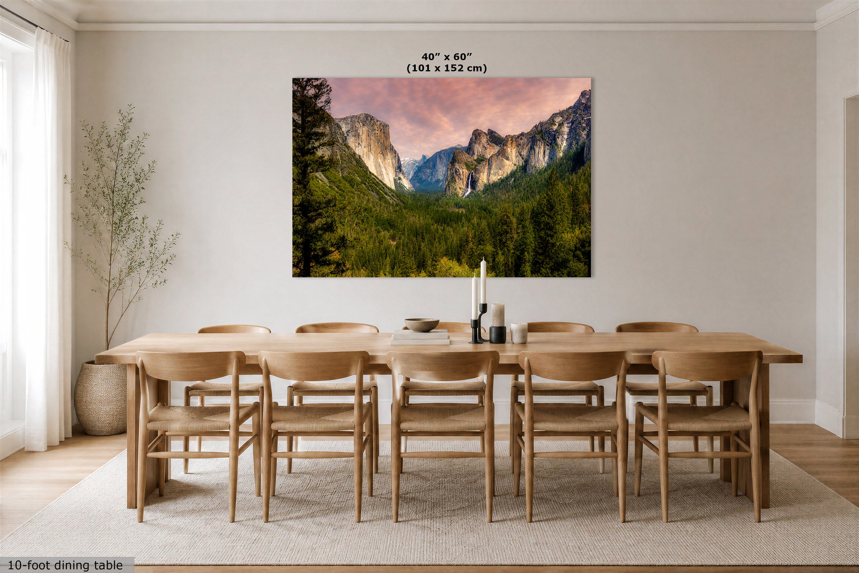 A large wall art print showing a mountain valley landscape with green trees and a pink sky, displayed above a wooden dining table and chairs in a bright dining room. Text at the top reads 40" x 60" (101 x 152 cm), and a small caption at the bottom left says 10-foot dining table.