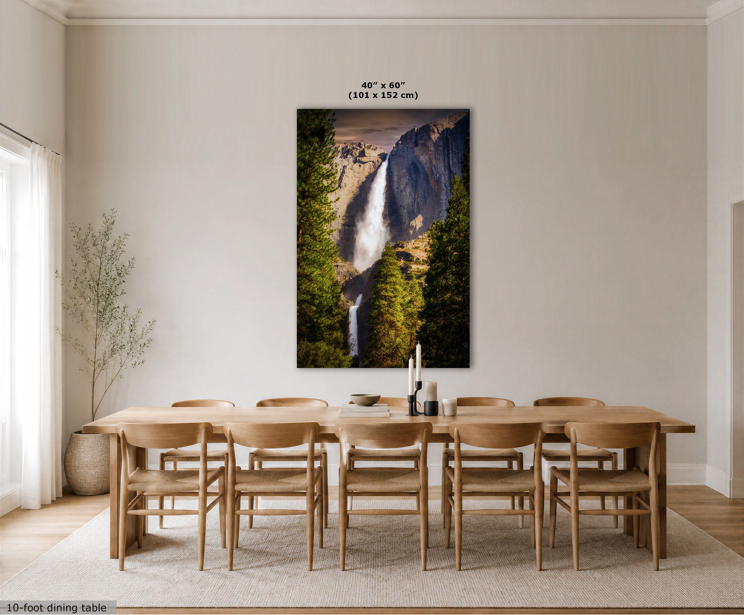A large vertical wall art print showing a waterfall in a forest canyon, displayed above a wooden dining table and chairs in a bright neutral dining room. The print includes size text at the top and a small text note in the lower left corner.