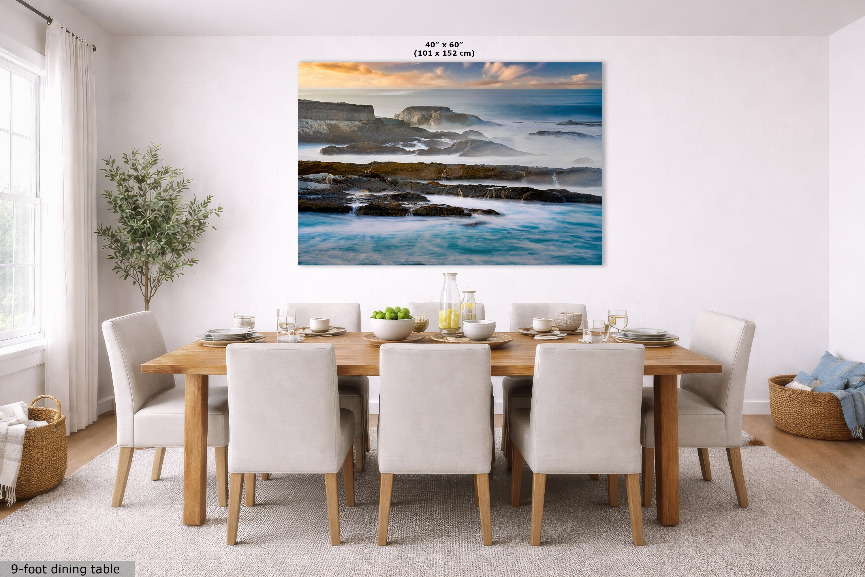 A large rectangular coastal landscape wall art print hangs above a 9-foot dining table with white upholstered chairs in a bright modern dining room. The artwork depicts blue ocean waves, dark rocky shoreline, and a warm sunset sky.