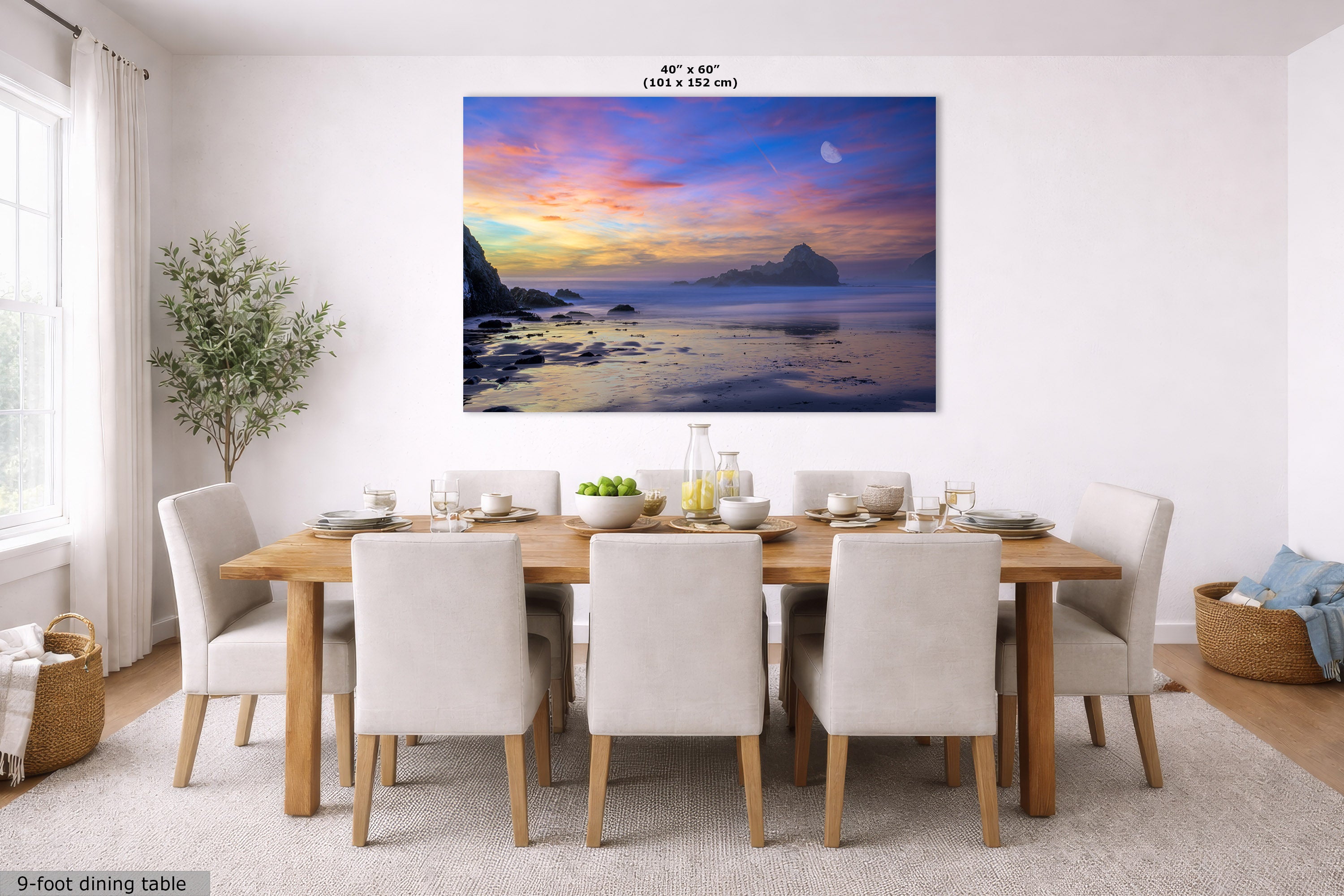 A large rectangular wall art print showing a colorful sunset over the ocean is displayed above a 9-foot dining table and six upholstered chairs in a bright, modern dining room. The print includes a small size label at the top reading 40 in x 60 in.