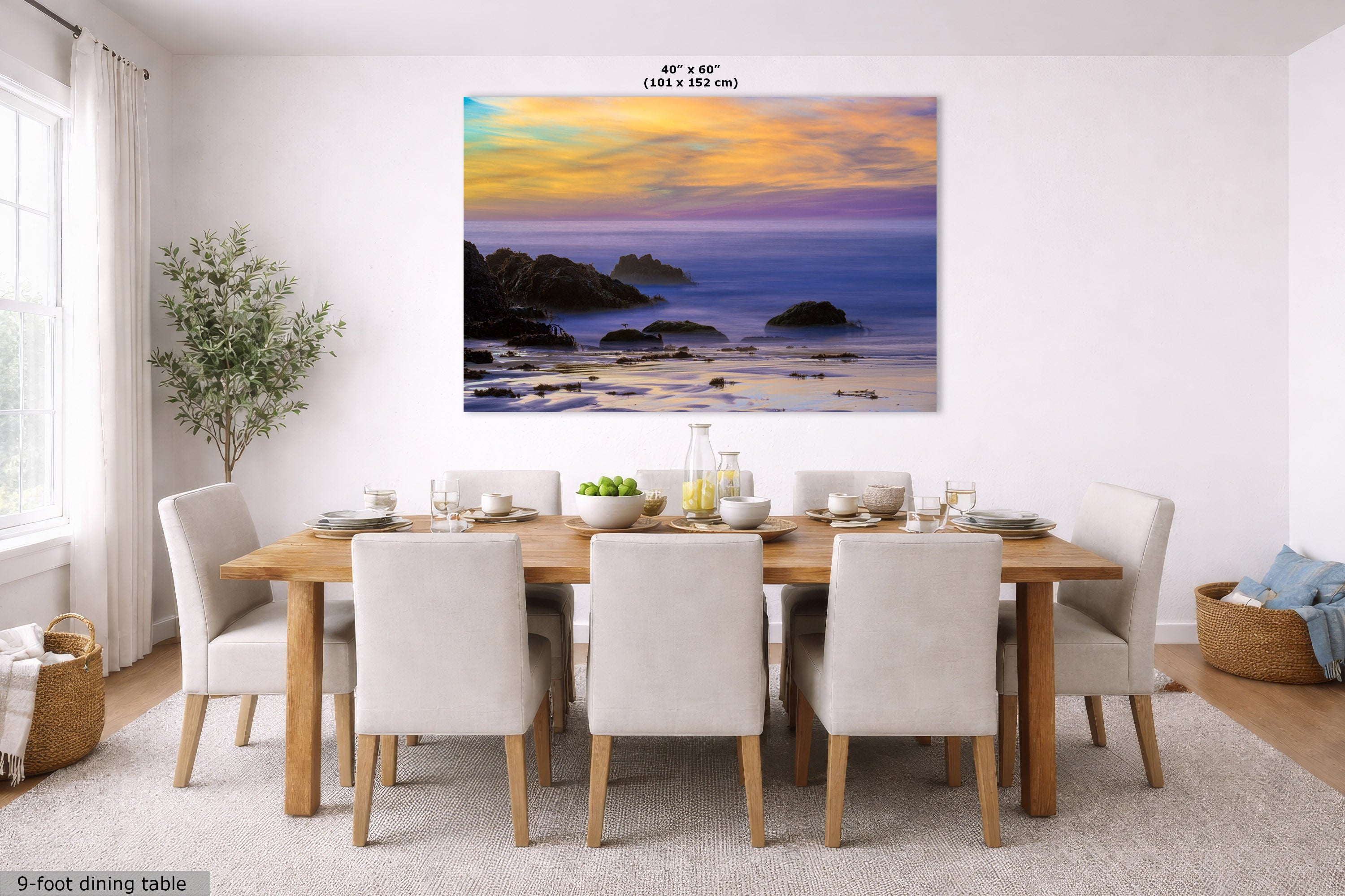 A large rectangular wall art print showing a coastal sunset scene with blue water, dark rocks, and warm orange-yellow sky, displayed above a dining table in a bright modern room. Small text at the top reads 40 in x 60 in (101 x 152 m), and text at bottom left says 9-foot dining table.