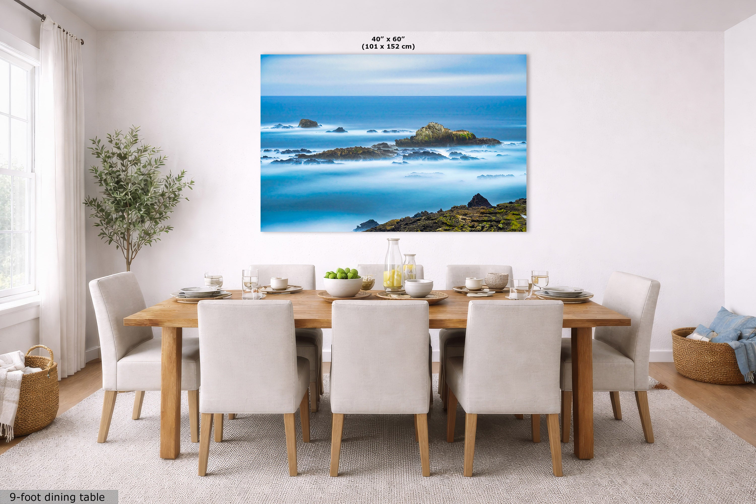 A large wall art print showing a coastal ocean scene with blue water, rocks, and green shoreline, displayed above a dining table in a bright modern room. Small text at the top reads 40 in x 60 in (101 x 152 cm), and text at the bottom left says 9-foot dining table.
