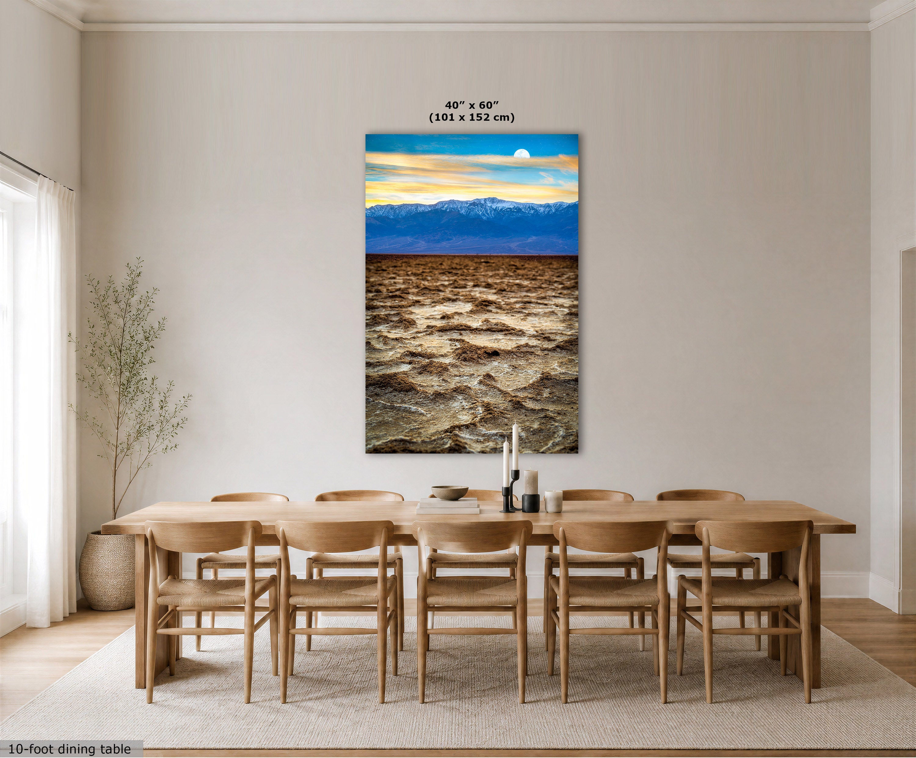 A large vertical canvas wall art print depicting a dramatic mountain and lake landscape, displayed in a modern dining room setting above a wooden dining table and chairs. The print features a blue sky, distant mountains, and a dark, textured lake in the foreground.