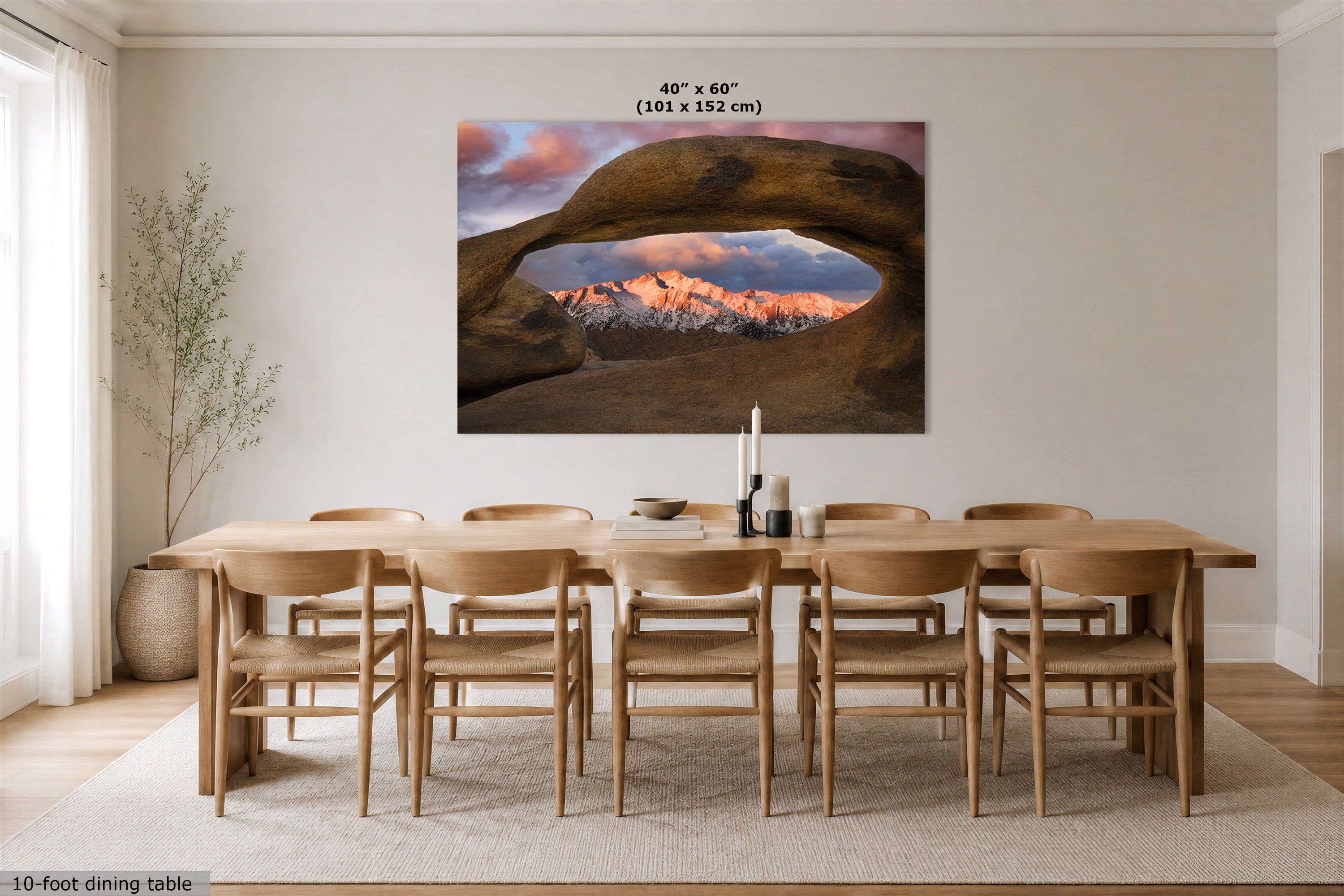 A large wall art print showing a desert rock arch framing a mountain sunset is displayed above a 10-foot dining table and chairs in a modern dining room. The print includes size text reading 40" x 60" (101 x 152 cm).