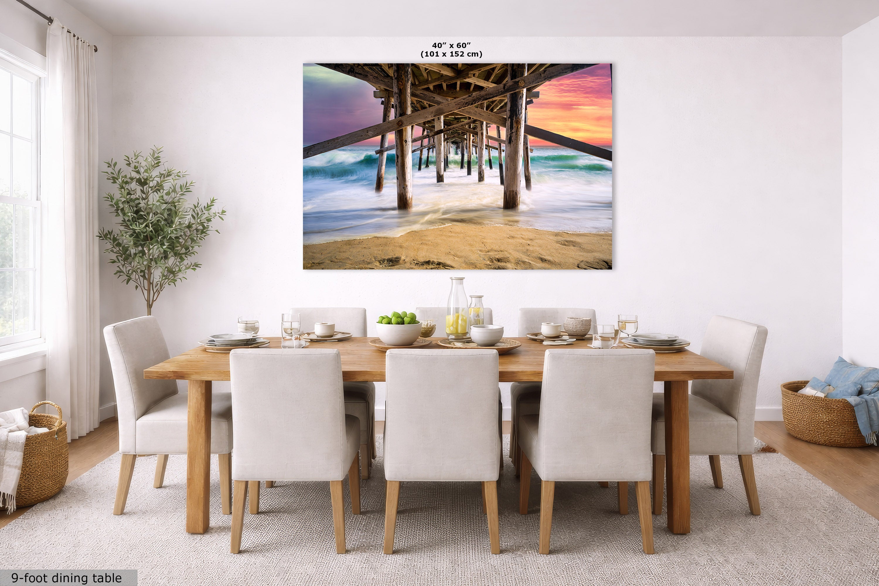 A large rectangular wall art print showing a pier over ocean waves at sunset, displayed above a dining table in a bright modern room. The print includes size text at the top and a small label at bottom left.