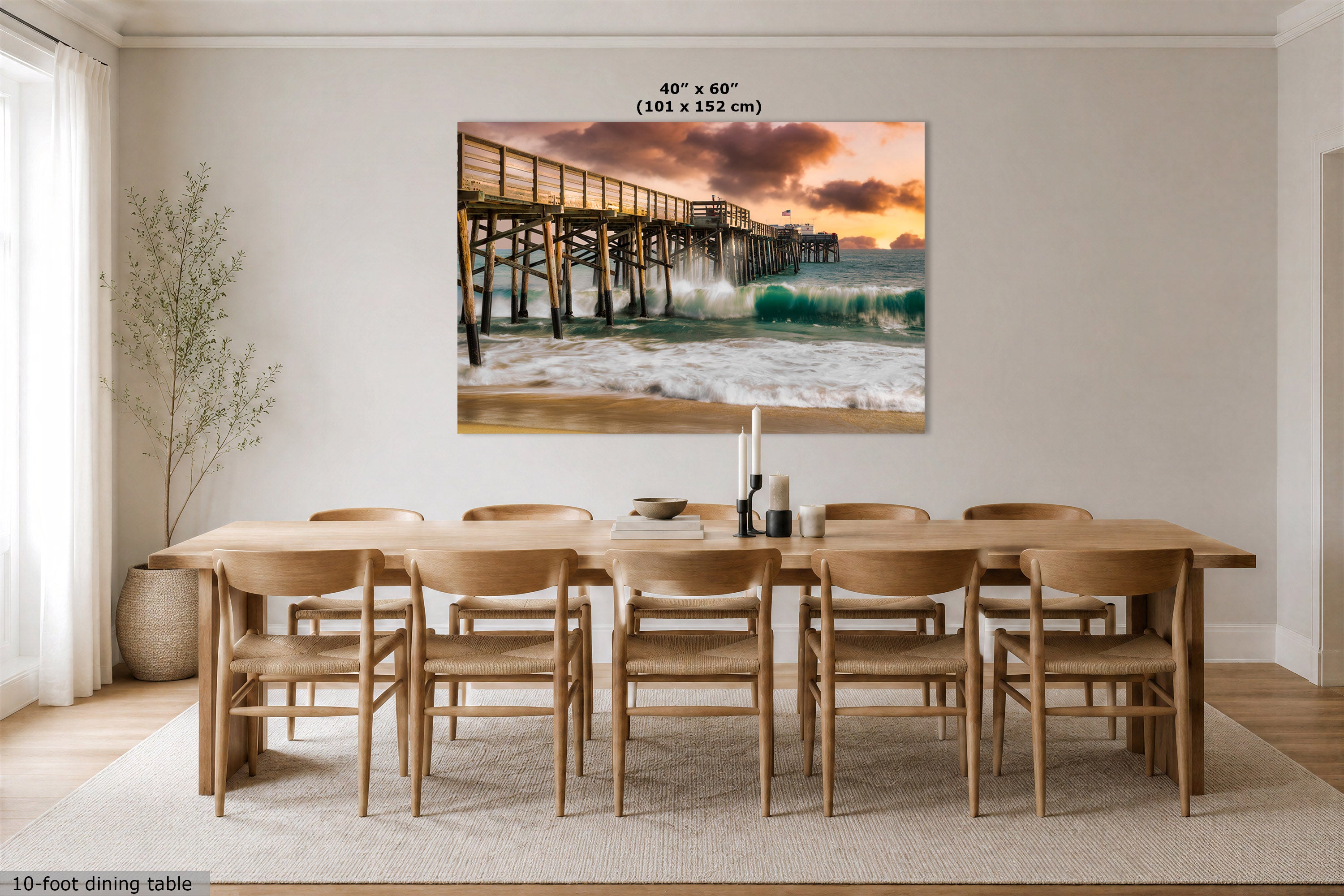 A large rectangular wall art print showing a pier over ocean waves at sunset, displayed above a wooden dining table and chairs in a bright dining room. Text at the top of the print reads 40" x 60" (101 x 152 cm).