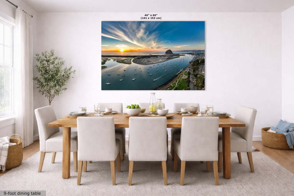 A large rectangular wall art print showing a sunset over a river with boats, displayed above a wooden dining table and chairs in a bright modern dining room. The print has dimensions labeled at the top, and a small caption in the lower left reads 9-foot dining table.