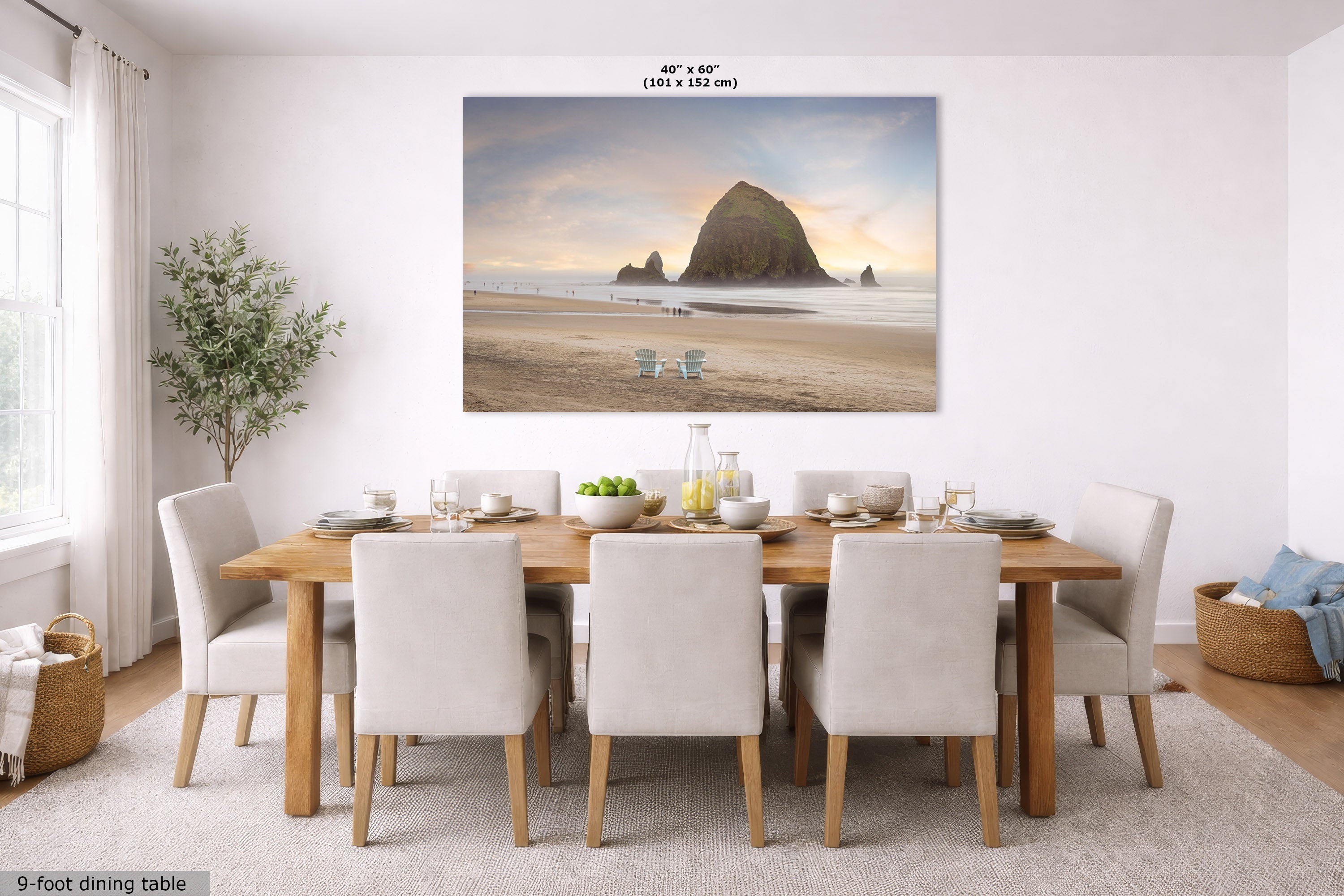 A large rectangular wall art print showing a beach scene with a large rock formation at sunset, displayed above a wooden dining table with white upholstered chairs in a bright dining room. Small text at the top reads 40 in x 60 in, and text at the bottom left reads 9-foot dining table.