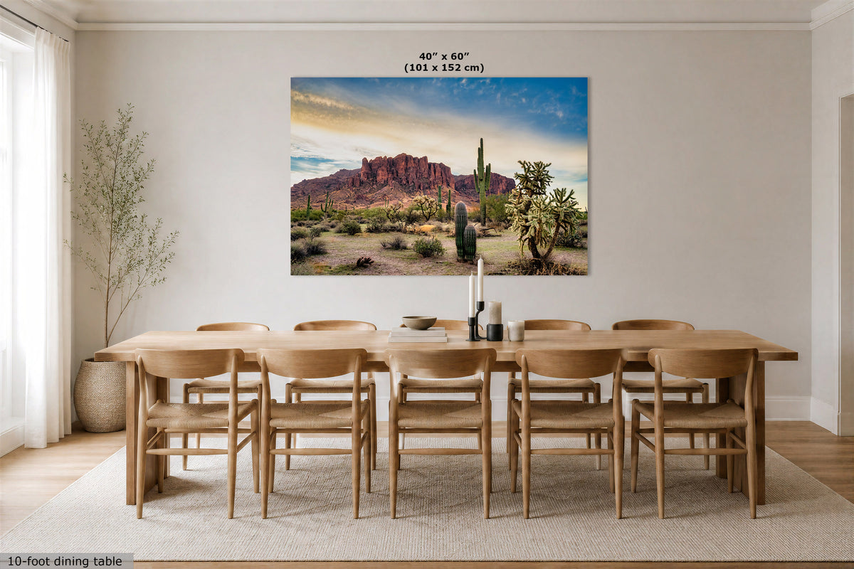A 40" x 60" canvas print of a desert landscape with red rock mountains, saguaro cacti, and a blue sky, displayed on a dining room wall above a wooden dining table and chairs.