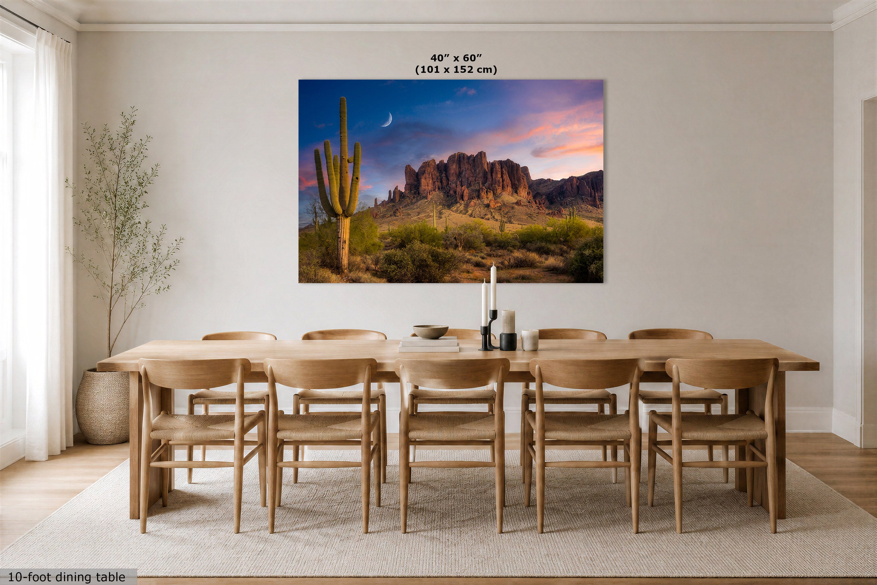 A large wall art print showing a desert landscape with a saguaro cactus, rocky mountains, and a colorful sunset sky, displayed above a wooden dining table and chairs in a bright dining room. Text at the top reads 48" x 60" (101 x 152 cm), and text at the bottom left says 10-foot dining table.
