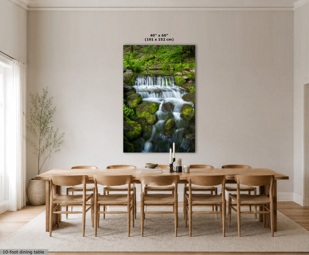 A large vertical wall art print showing a lush green waterfall over mossy rocks, displayed above a light wood dining table and chairs in a bright, modern dining room. The print has a small size label at the top reading 40" x 60" (101 x 152 cm).