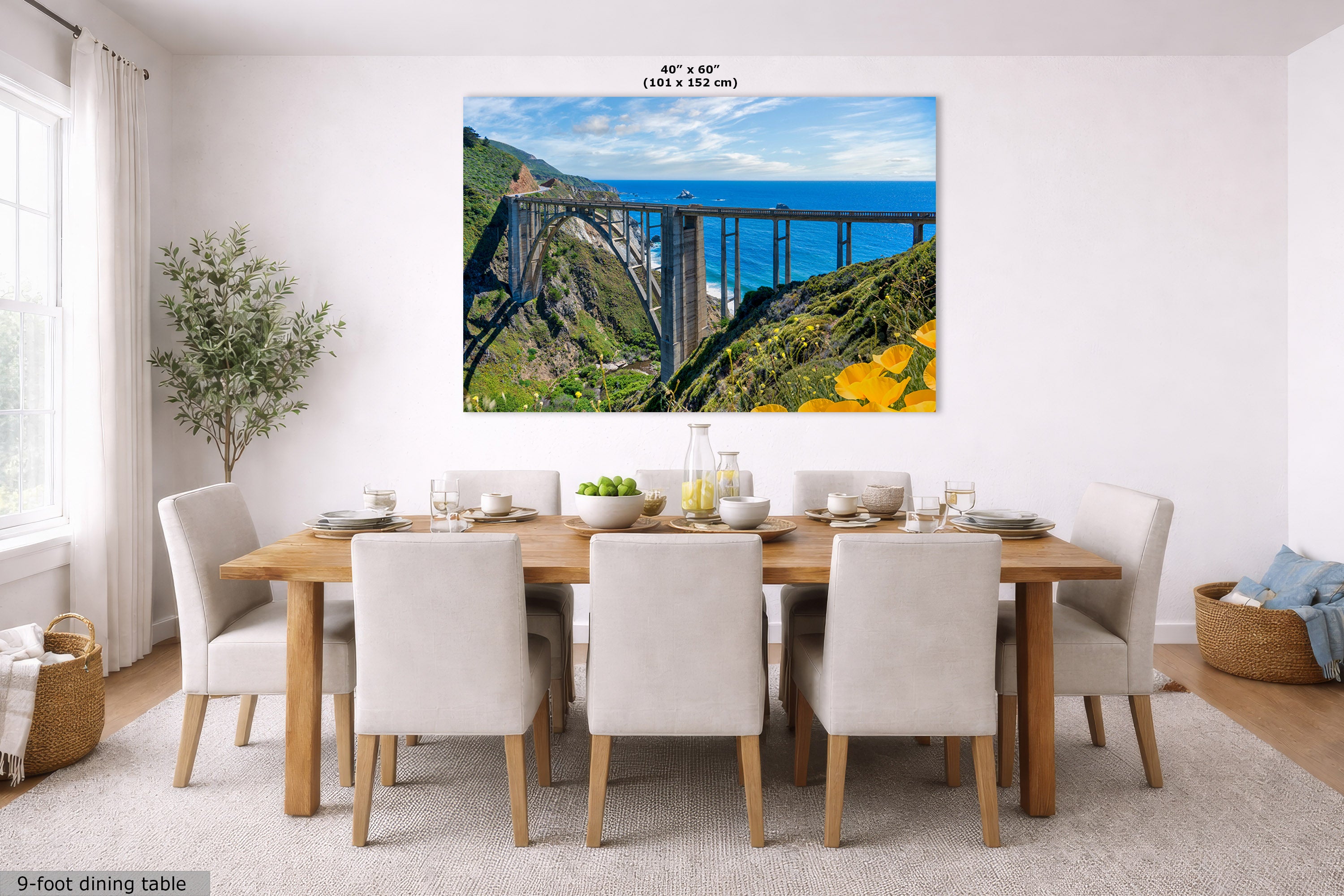 A large rectangular wall art print showing a coastal bridge over green cliffs and blue ocean, displayed above a dining table in a bright modern room. Small text at the top reads 40 x 60 inches, and text at the bottom left says 9-foot dining table.