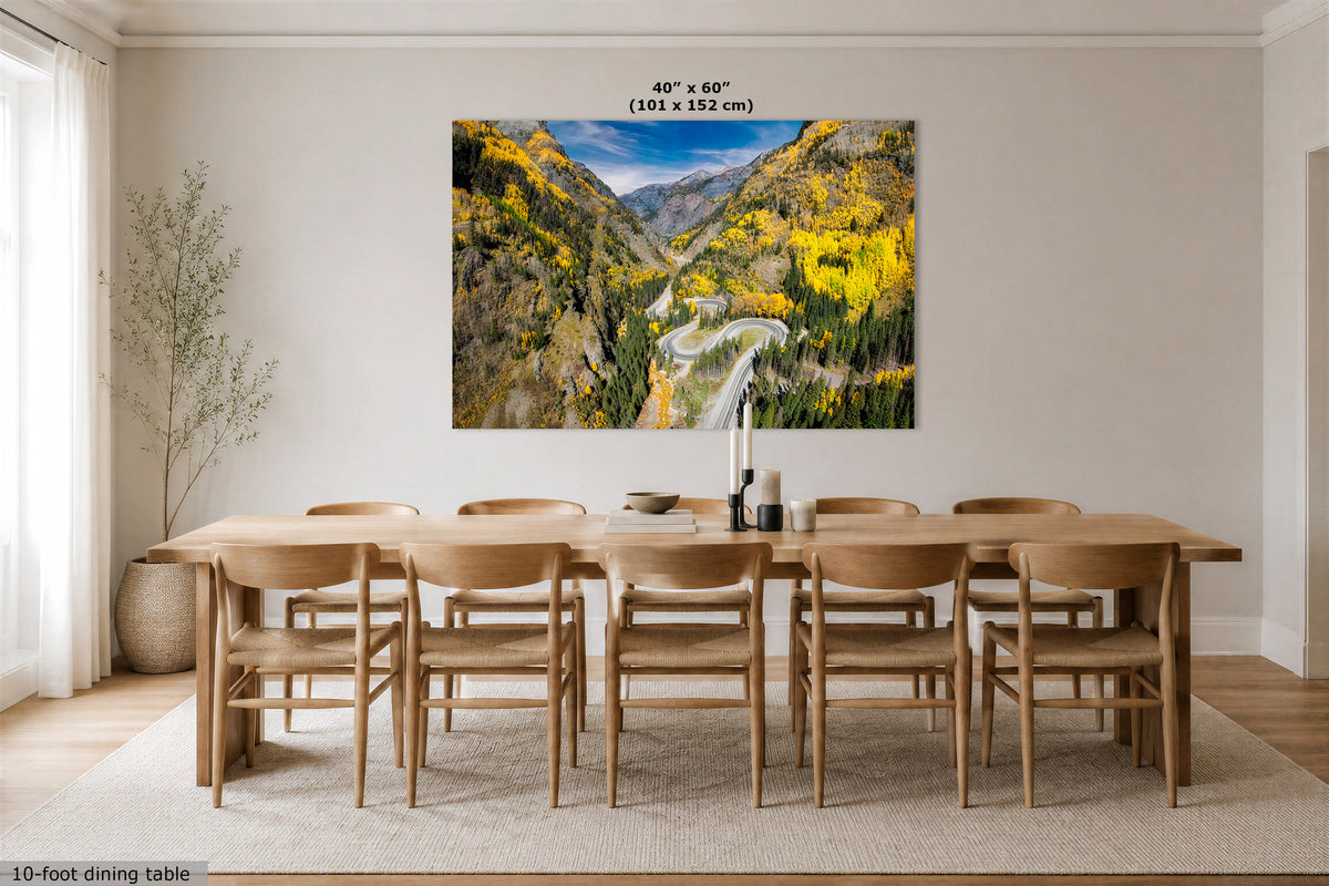 A large wall art print showing a mountain valley with yellow foliage and a winding road, displayed above a wooden dining table and chairs in a bright dining room. Text at the top reads 40" x 60" (101 x 152 cm), and a small caption at the bottom left says 10-foot dining table.