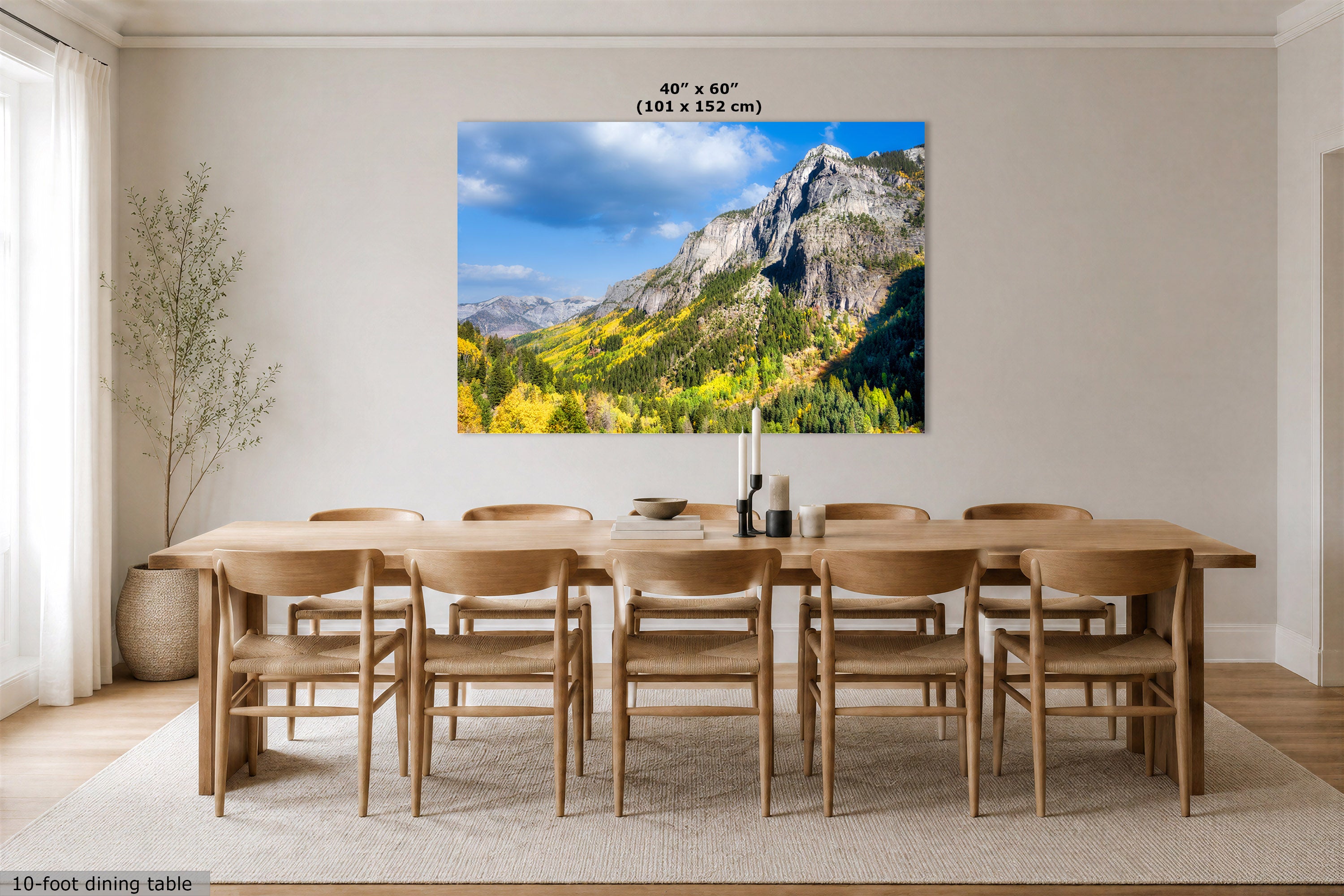 A large wall art print showing a mountain landscape with blue sky and yellow-green foliage, displayed above a wooden dining table and chairs in a bright dining room. Text at the top reads 40" x 60" (101 x 152 cm), and a small caption at the bottom left says 10-foot dining table.