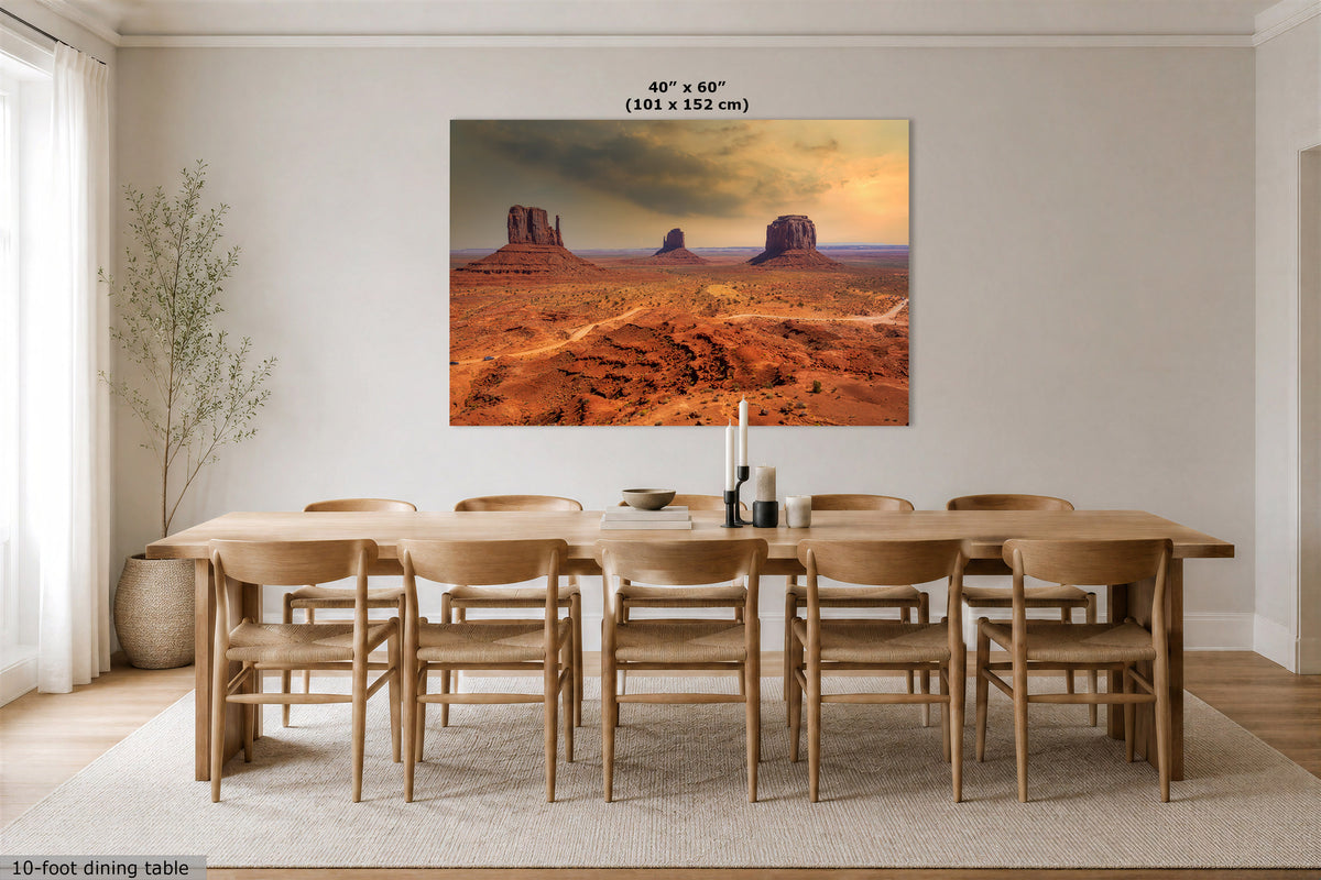 A large rectangular wall art print showing a desert landscape with red rock formations under a dramatic sky, displayed above a wooden dining table in a bright neutral dining room. Text at the top reads 40" x 60" (101 x 152 cm).