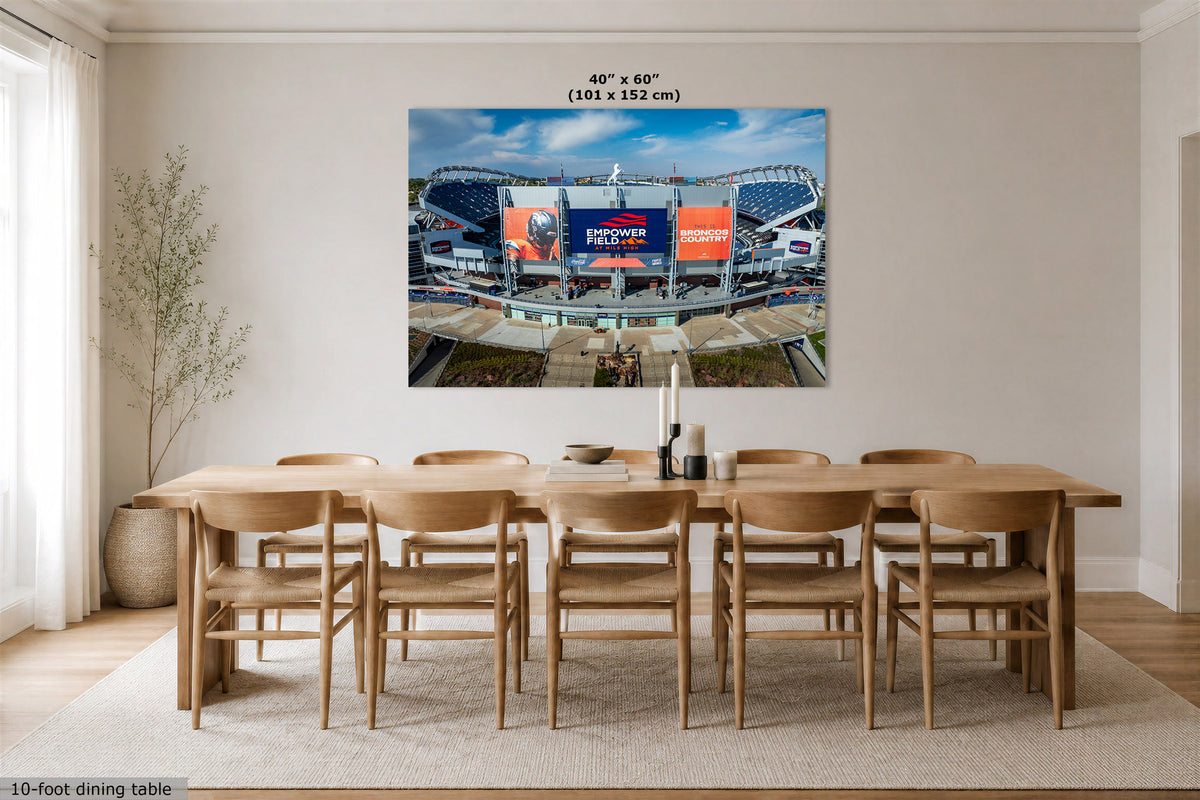 A large rectangular wall art print showing a stadium scene with a large screen reading 'BRONCOS COUNTRY', displayed above a wooden dining table and chairs in a bright modern dining room. Text at the top indicates the print size as 40" x 60" (101 x 152 cm).
