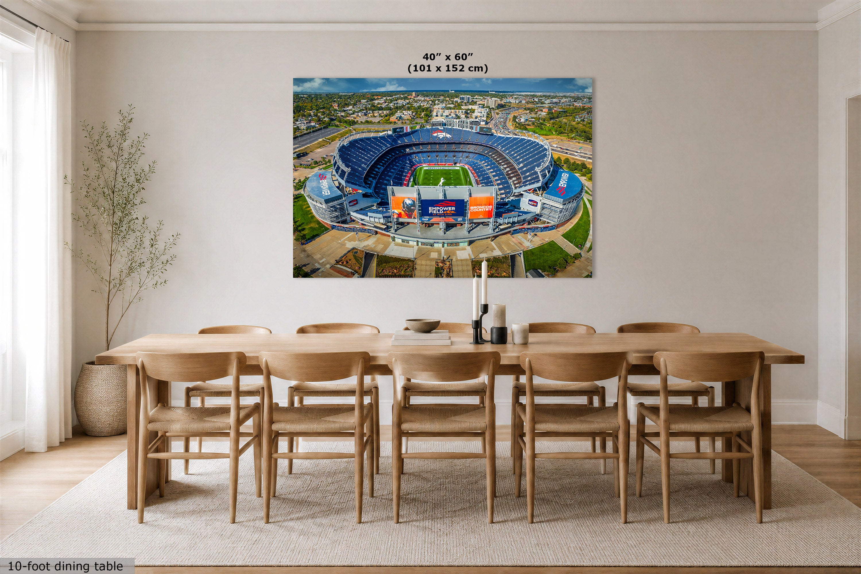 A Denver Broncos Stadium art print showing an aerial view of Mile High Stadium, displayed above a wooden dining table in a bright modern room. The print includes size text at the top reading 40" x 60" (101 x 152 cm).