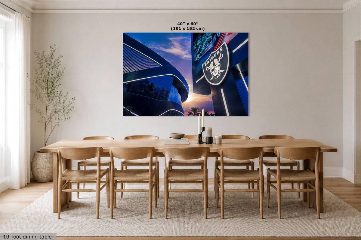 A large rectangular wall art print showing a blue and black stadium scene with the Raiders logo, displayed above a wooden dining table and chairs in a bright modern room. Text at the top reads 40" x 60" (101 x 152 cm), and text at the bottom left says 10-foot dining table.