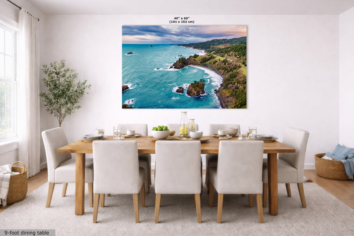 A large rectangular coastal landscape wall art print hangs above a wooden dining table with white upholstered chairs in a bright modern dining room. The artwork shows turquoise ocean water, rocky coastline, green hills, and a dramatic cloudy sky.