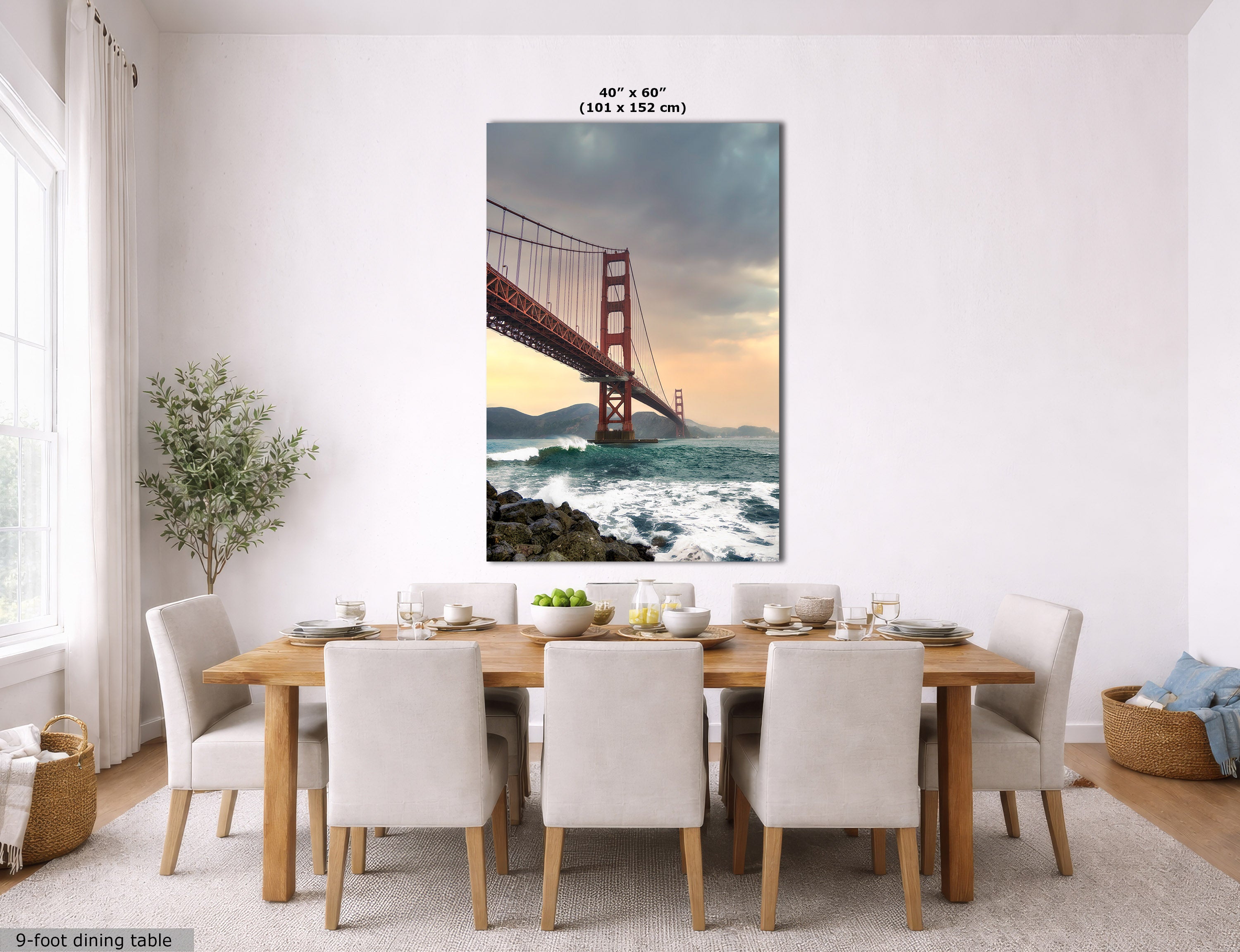 A large vertical wall art print of the Golden Gate Bridge over ocean waves hangs above a wooden dining table with upholstered chairs in a bright, modern dining room. The print includes size text at the top and a small note at bottom left.