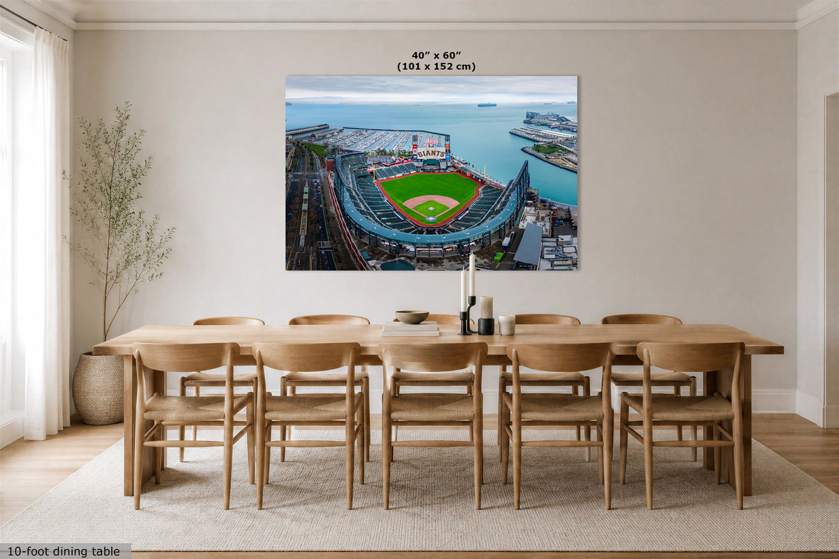 A large wall art print showing an aerial view of a baseball stadium by the water, displayed above a wooden dining table and chairs in a bright modern room. The print includes size text at the top reading 40" x 60" (101 x 152 cm).