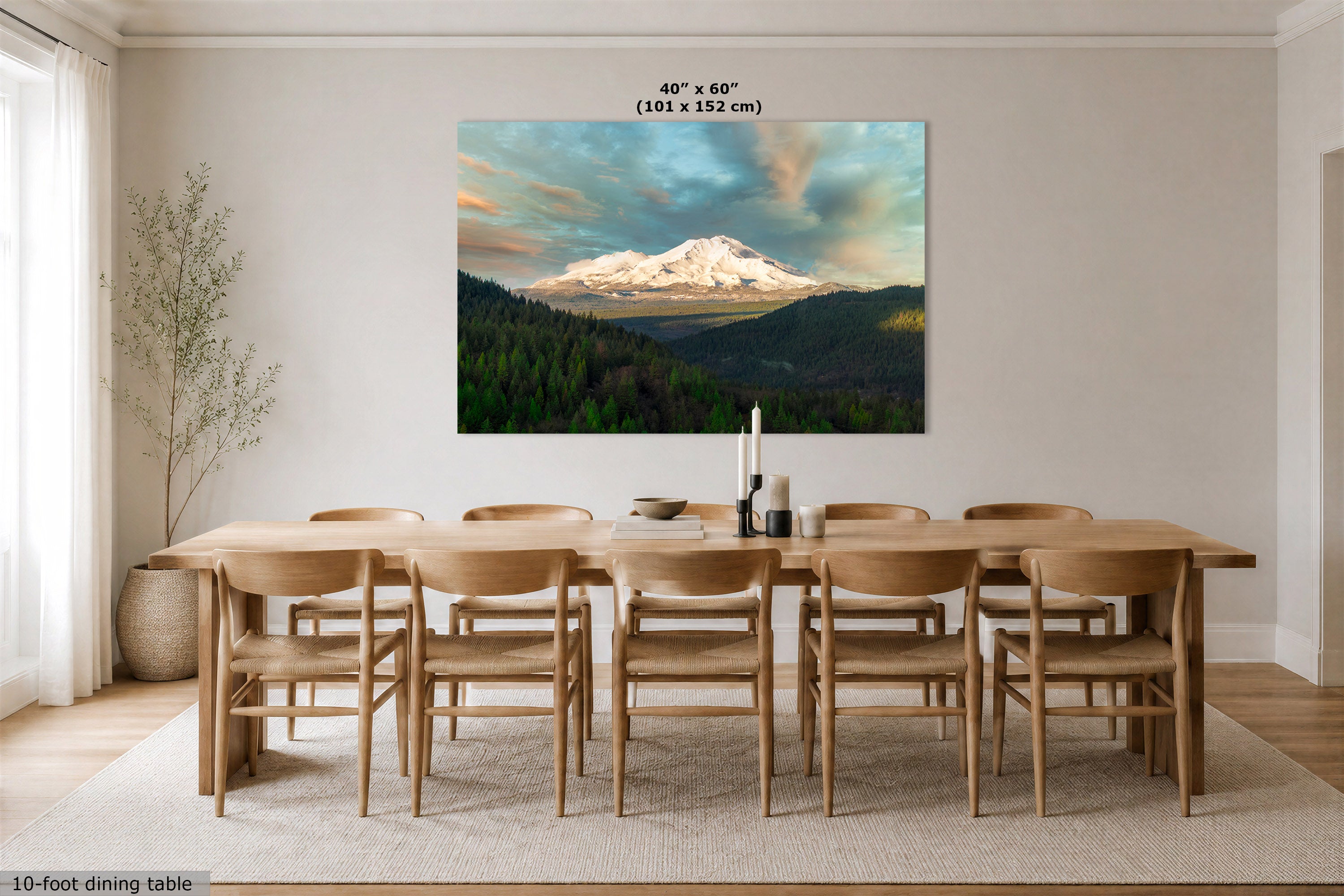 A large rectangular wall art print showing a snow-capped mountain landscape with green forest below, displayed above a wooden dining table and chairs in a bright dining room. The print includes size text reading 40 x 60 in (101 x 152 cm).