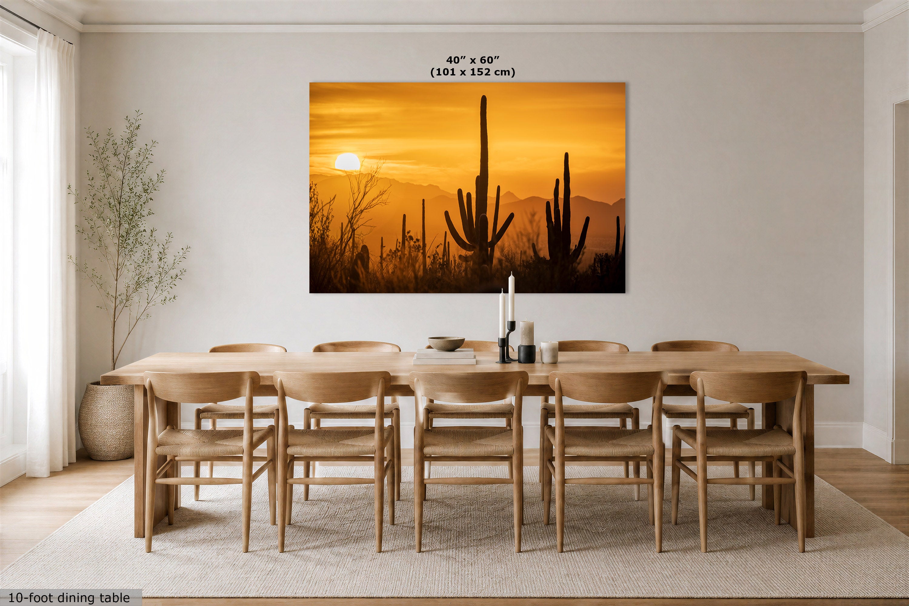 A large rectangular wall art print showing a desert sunset with saguaro cacti, displayed above a wooden dining table and chairs in a bright dining room. The print includes size text at the top and a small note at the bottom left.