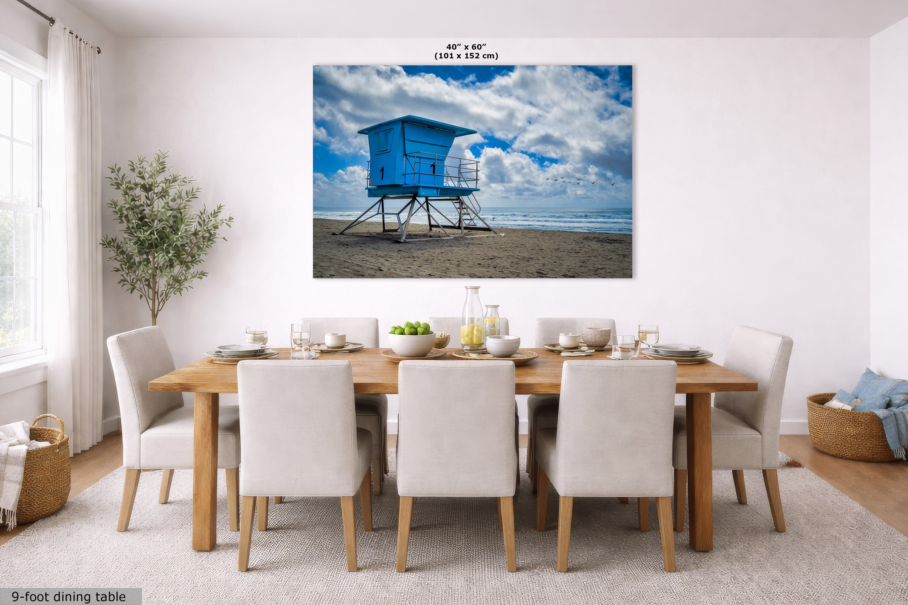 A large square wall art print showing a blue lifeguard tower on a sandy beach under a cloudy blue sky, displayed above a wooden dining table with white chairs in a bright modern dining room. Small text at the top reads 40" x 60" and at the bottom left says 9-foot dining table.