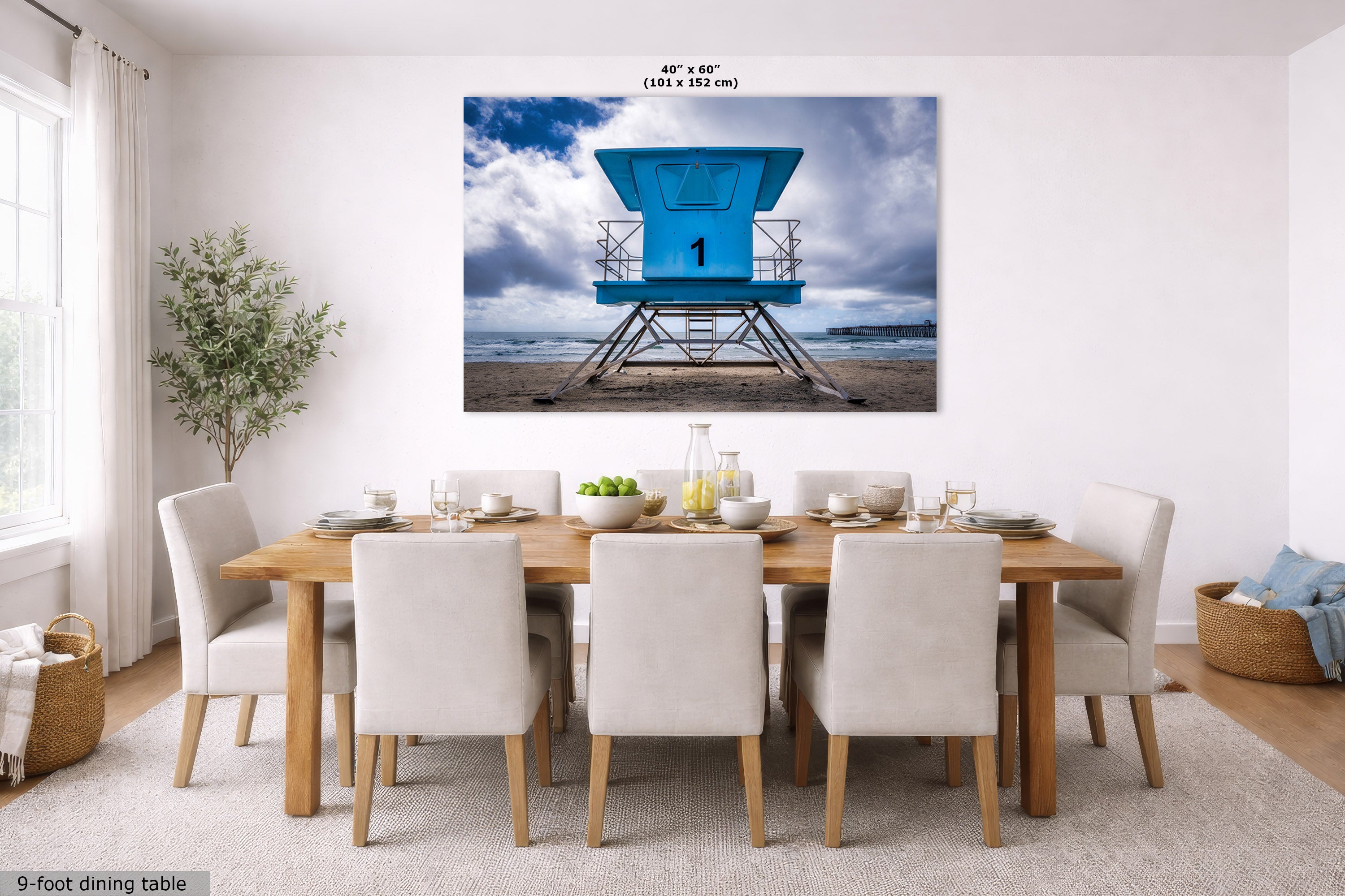 A large wall art print showing a bright blue lifeguard tower on a beach under a cloudy sky, displayed above a wooden dining table with white chairs in a modern dining room. The print includes size text at the top and a small label at the bottom left.