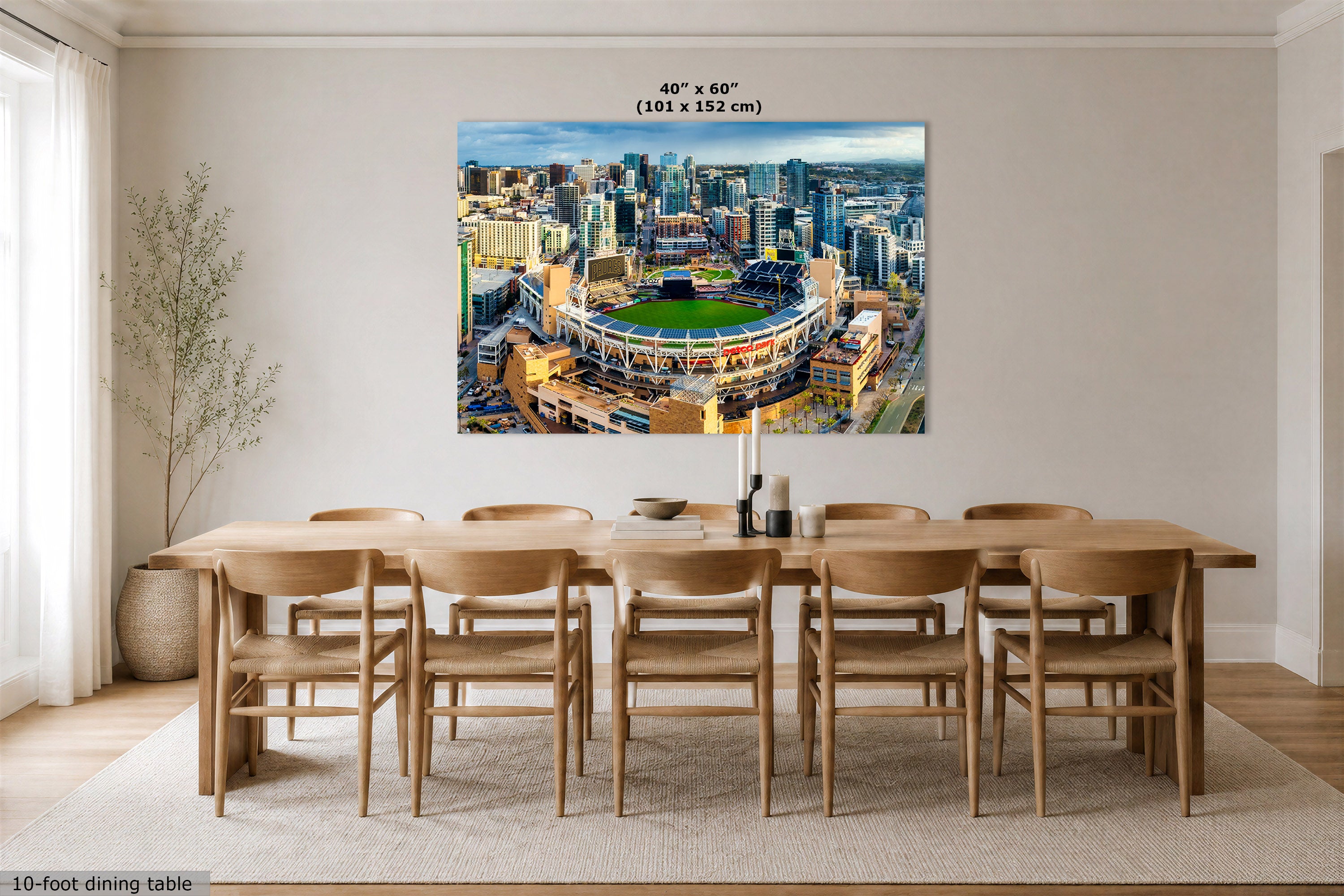 A large wall art print showing an aerial view of a stadium and city skyline, displayed above a wooden dining table in a bright modern dining room. Text at the top reads 40" x 60" (101 x 152 cm), and text at the bottom left says 10-foot dining table.