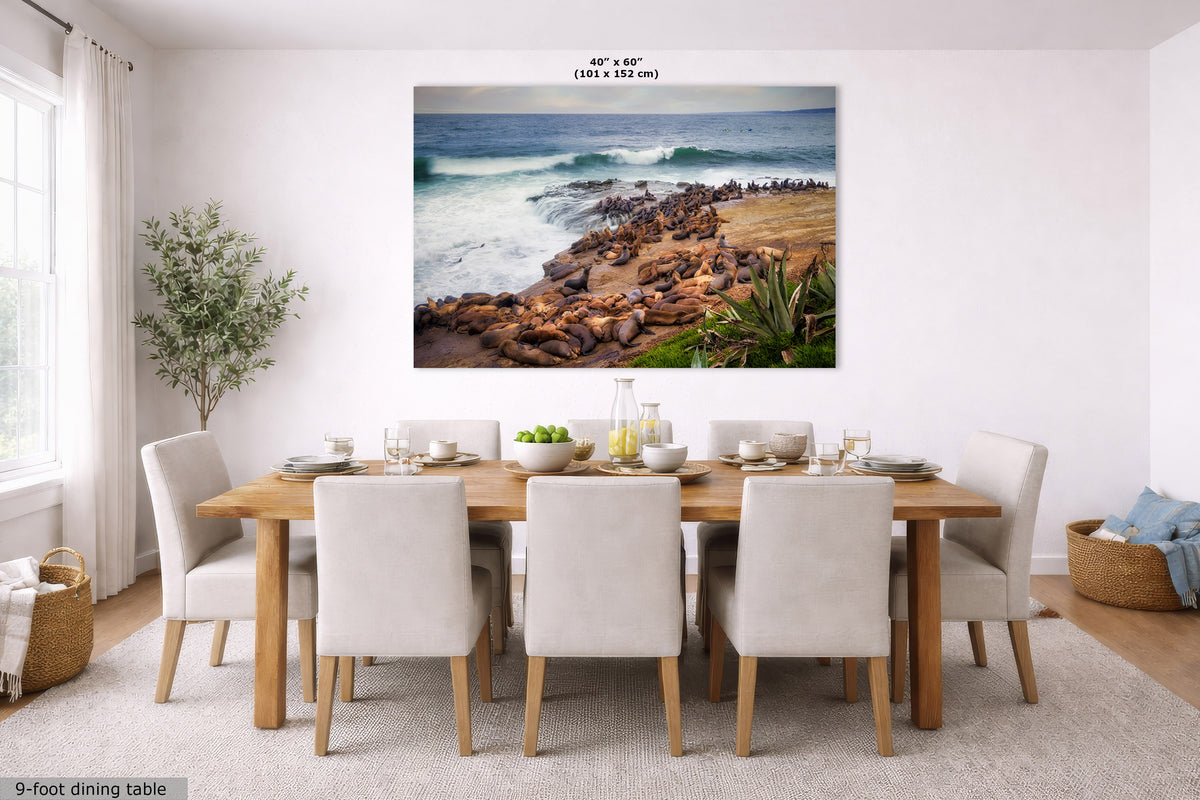 A large framed wall art print showing a rocky beach and ocean scene, displayed above a wooden dining table with white upholstered chairs in a bright modern dining room. The print includes a small size label reading 40" x 60" (101 x 152 cm).