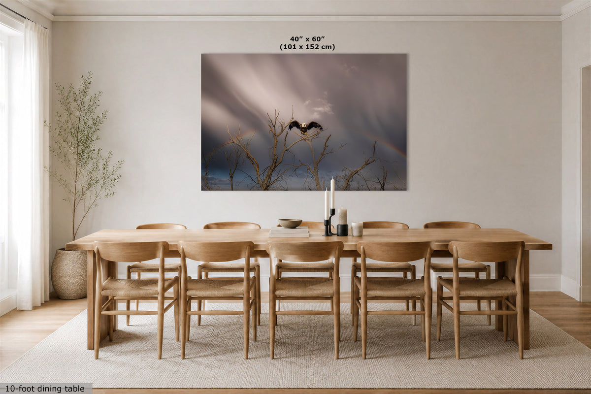 A large rectangular wall art print showing a stormy sky with two birds on bare branches, displayed above a wooden dining table and chairs in a bright neutral dining room. The print includes size text at the top and a small text credit at the bottom left.