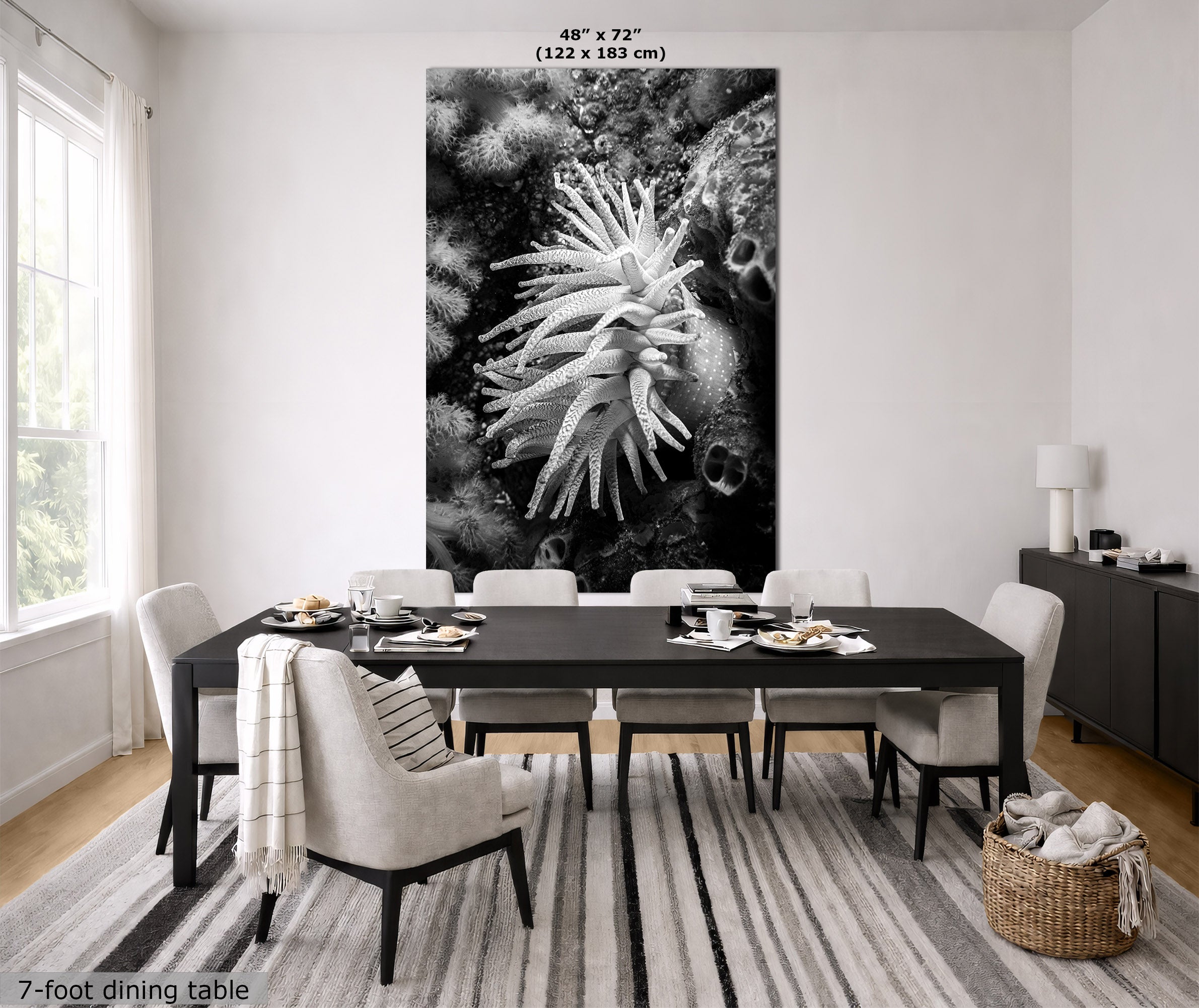A large black and white wall art print of an anemone on a coral reef, shown hanging in a modern dining room above a dark wood dining table and chairs. The print includes size text at the top and a note at the bottom indicating the 7-foot dining table scale.